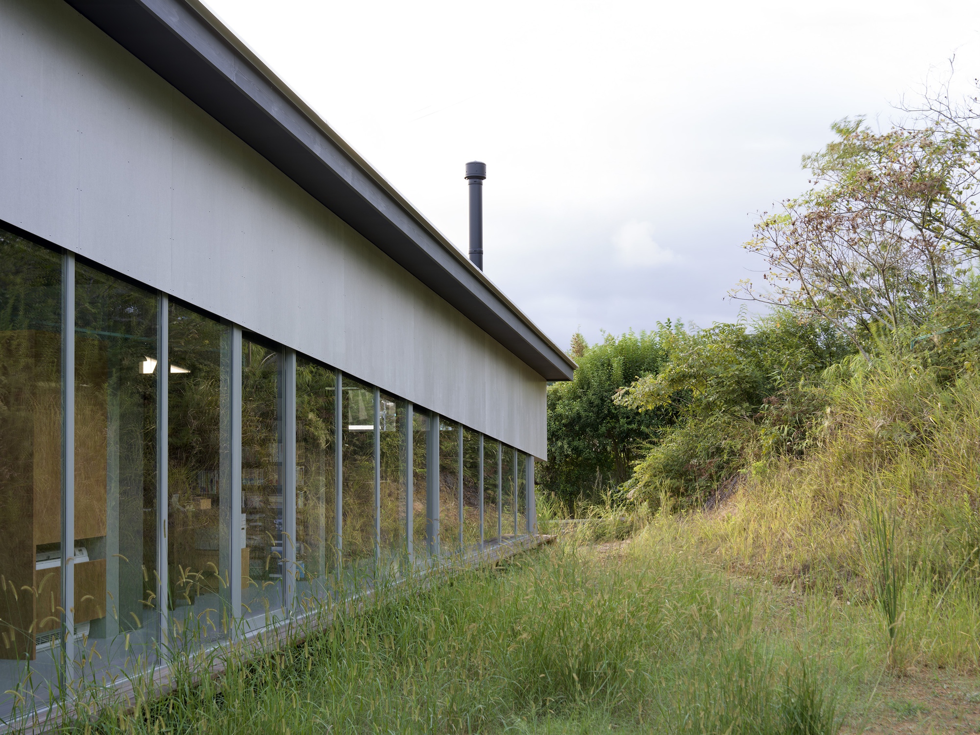 Long glazed wall with narrow vertical framing set into a sloping meadow under overcast skies