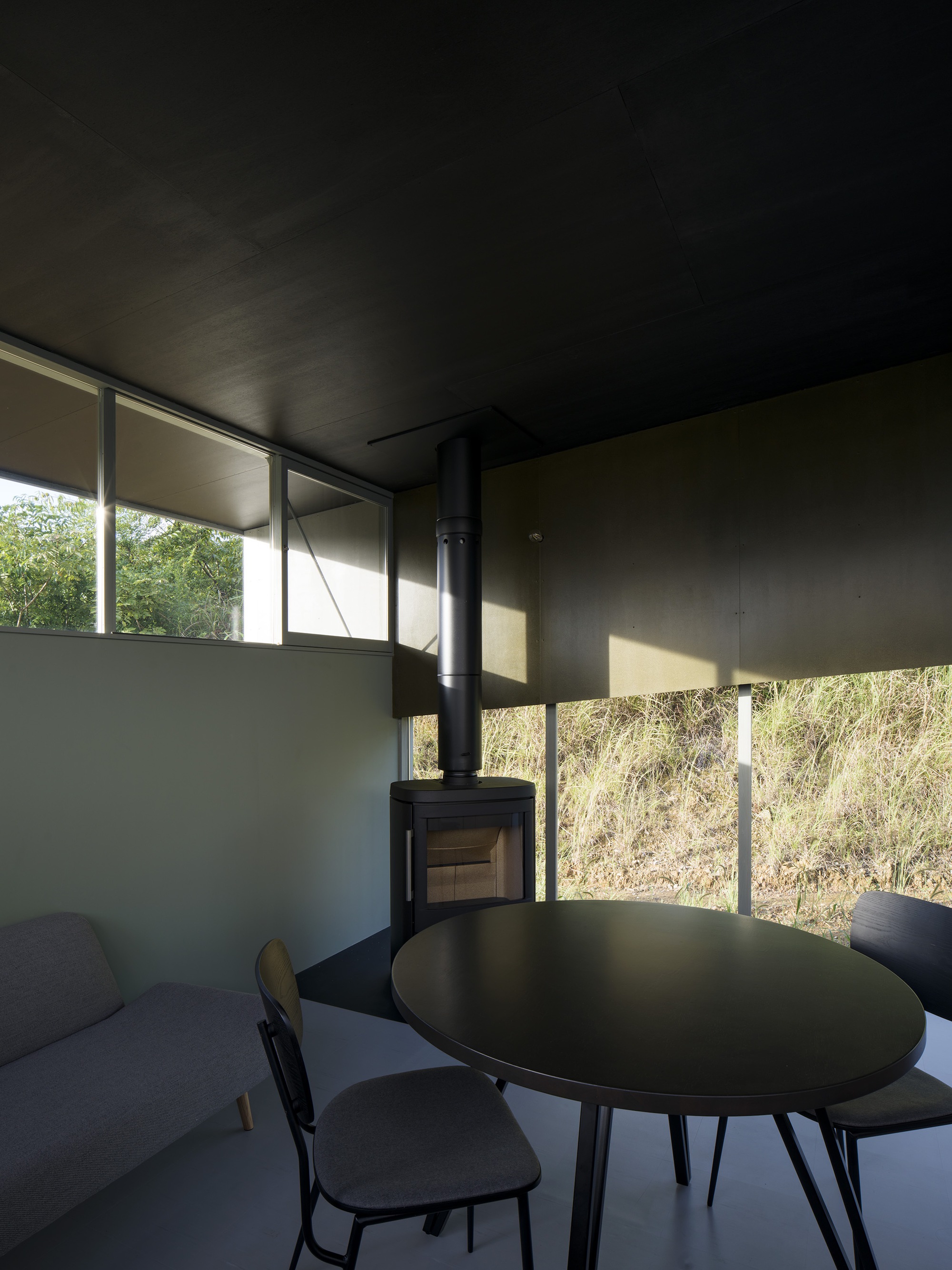 Living space with freestanding wood stove and horizontal ribbon windows framing hillside vegetation