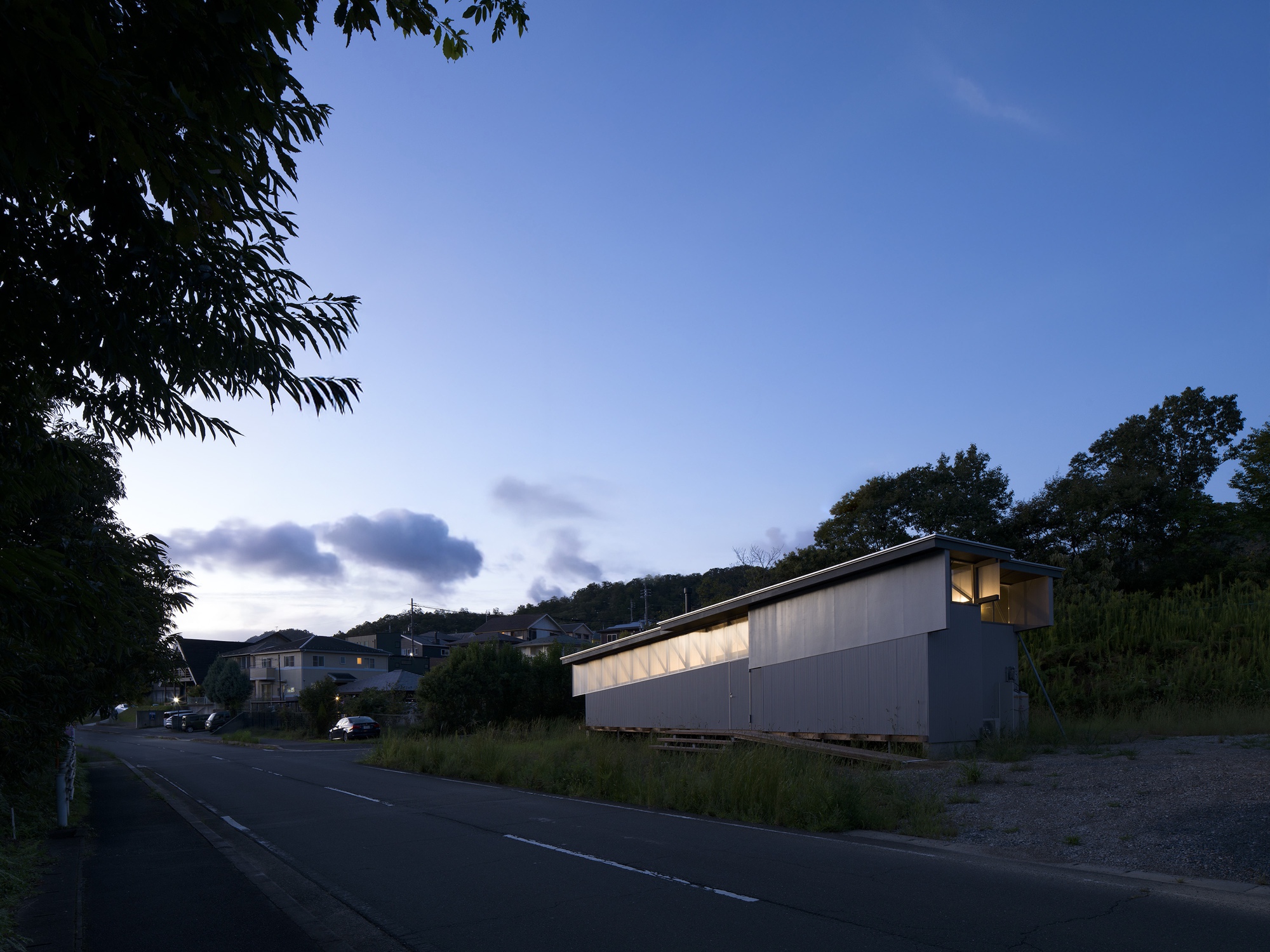 Street view of the residence with illuminated clerestory windows at dusk beneath wooded hillside