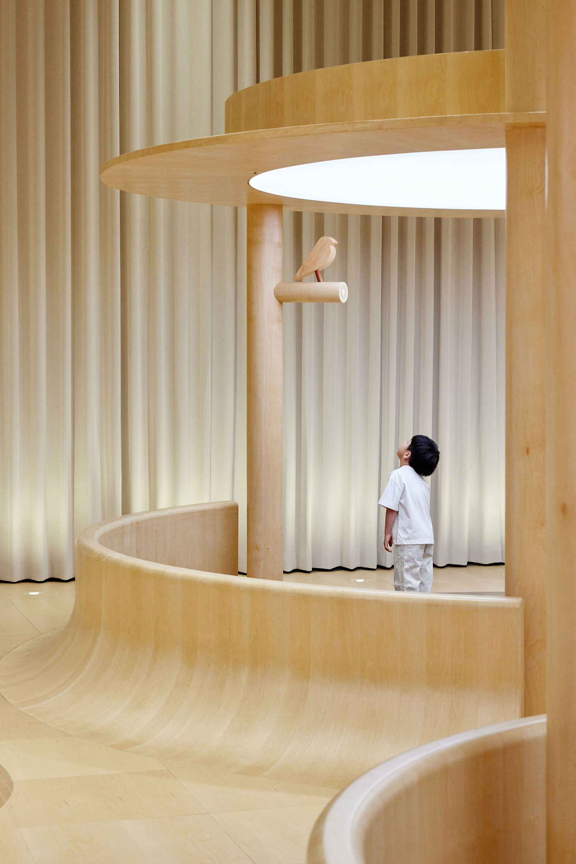 Multilevel plywood platforms with vertical curtain panels and a child standing beneath a perch