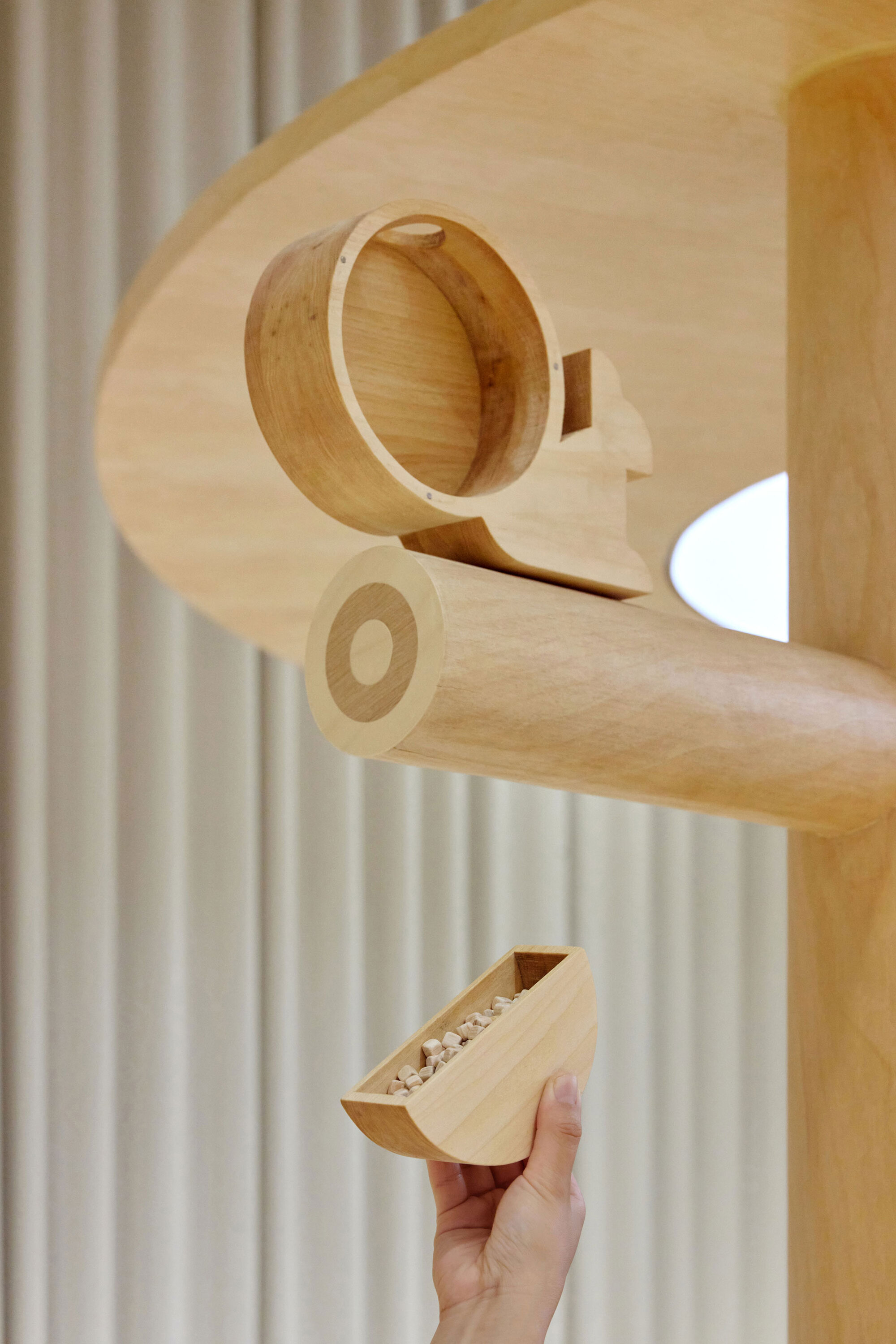 Hand holding a wooden toy piece beneath a cylindrical timber drum suspended from plywood structure