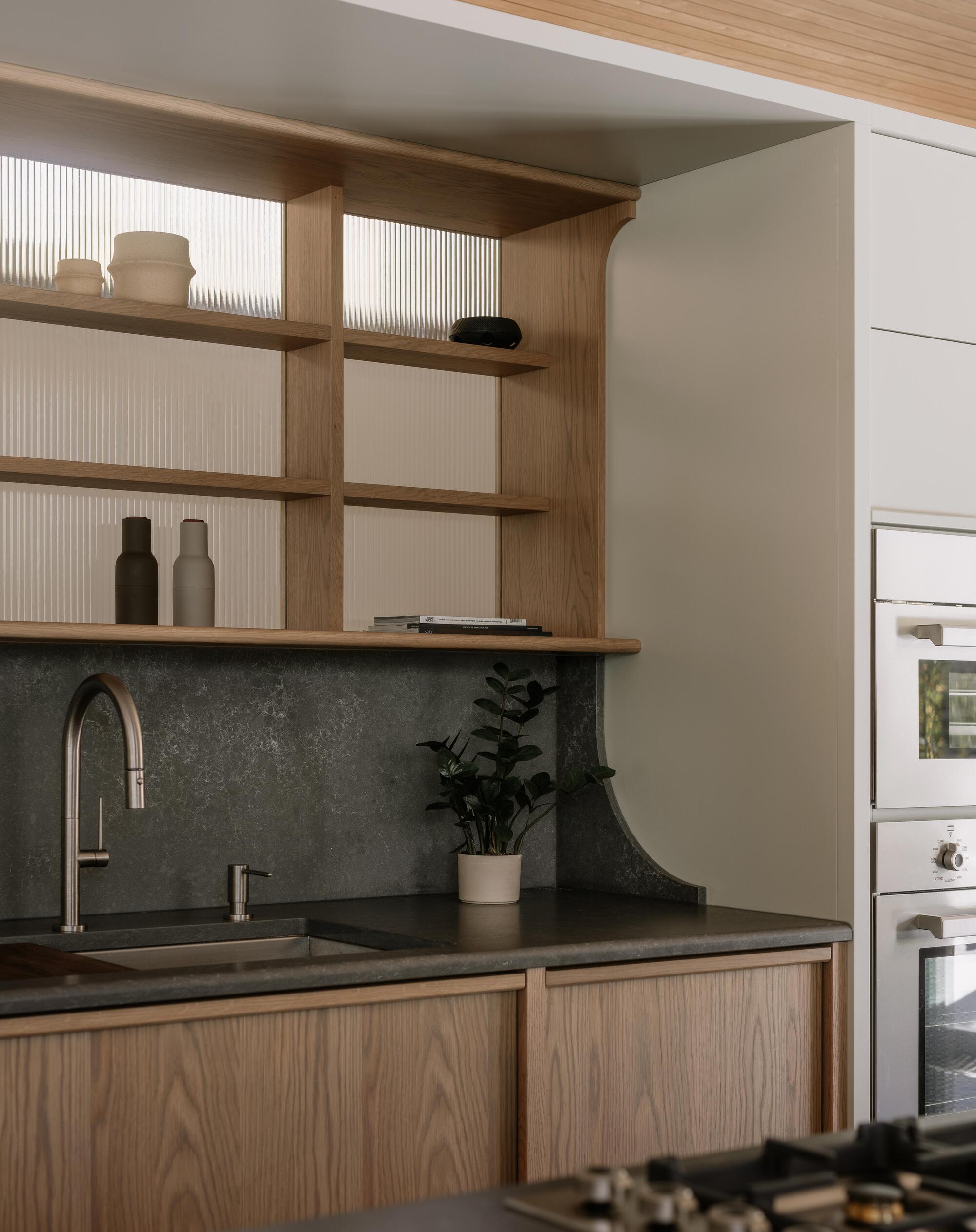 Kitchen counter with timber shelving, fluted glass panels, and dark stone backsplash under natural light