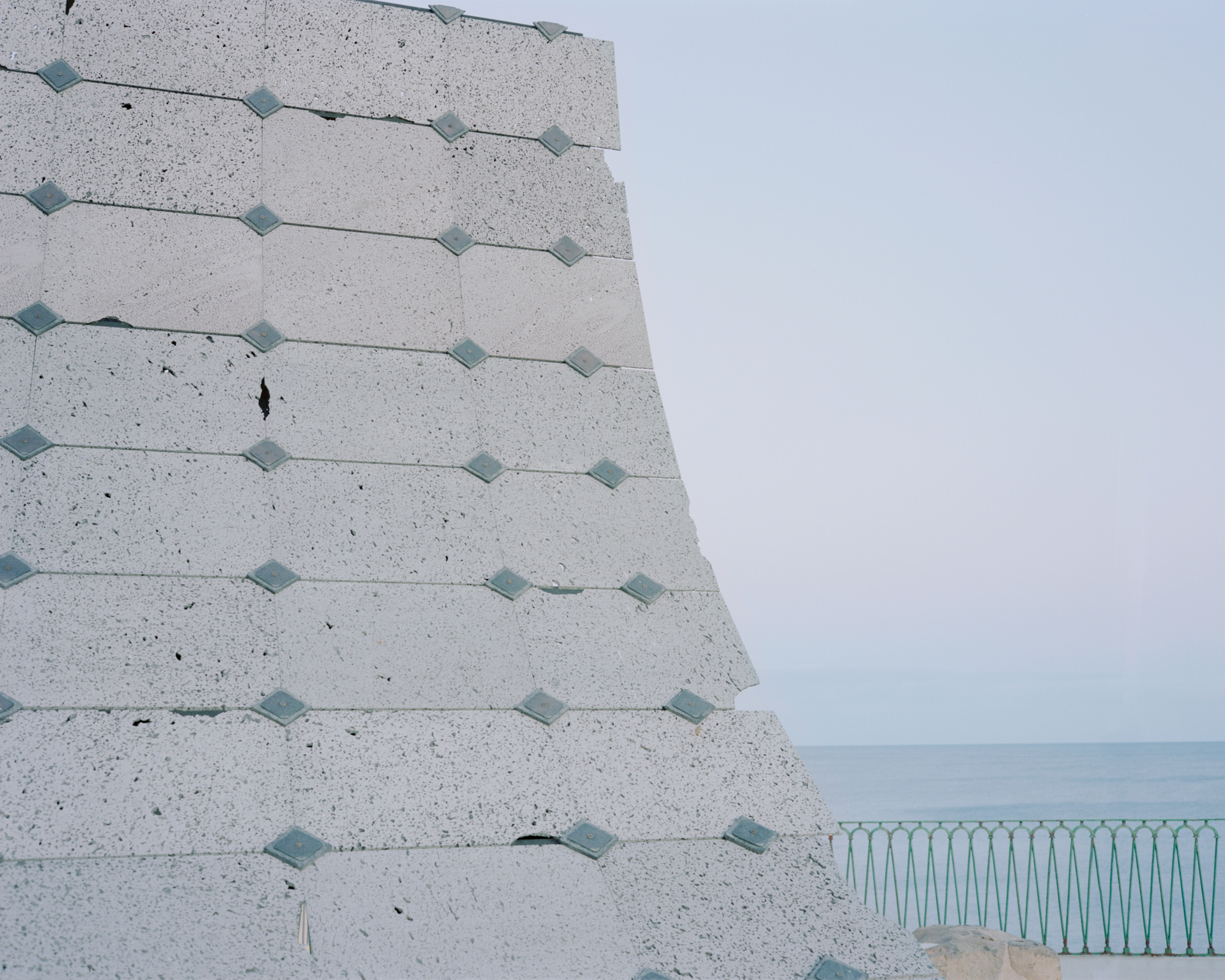 Corner of the tilted concrete wall with metal fixings overlooking a pale blue sea horizon