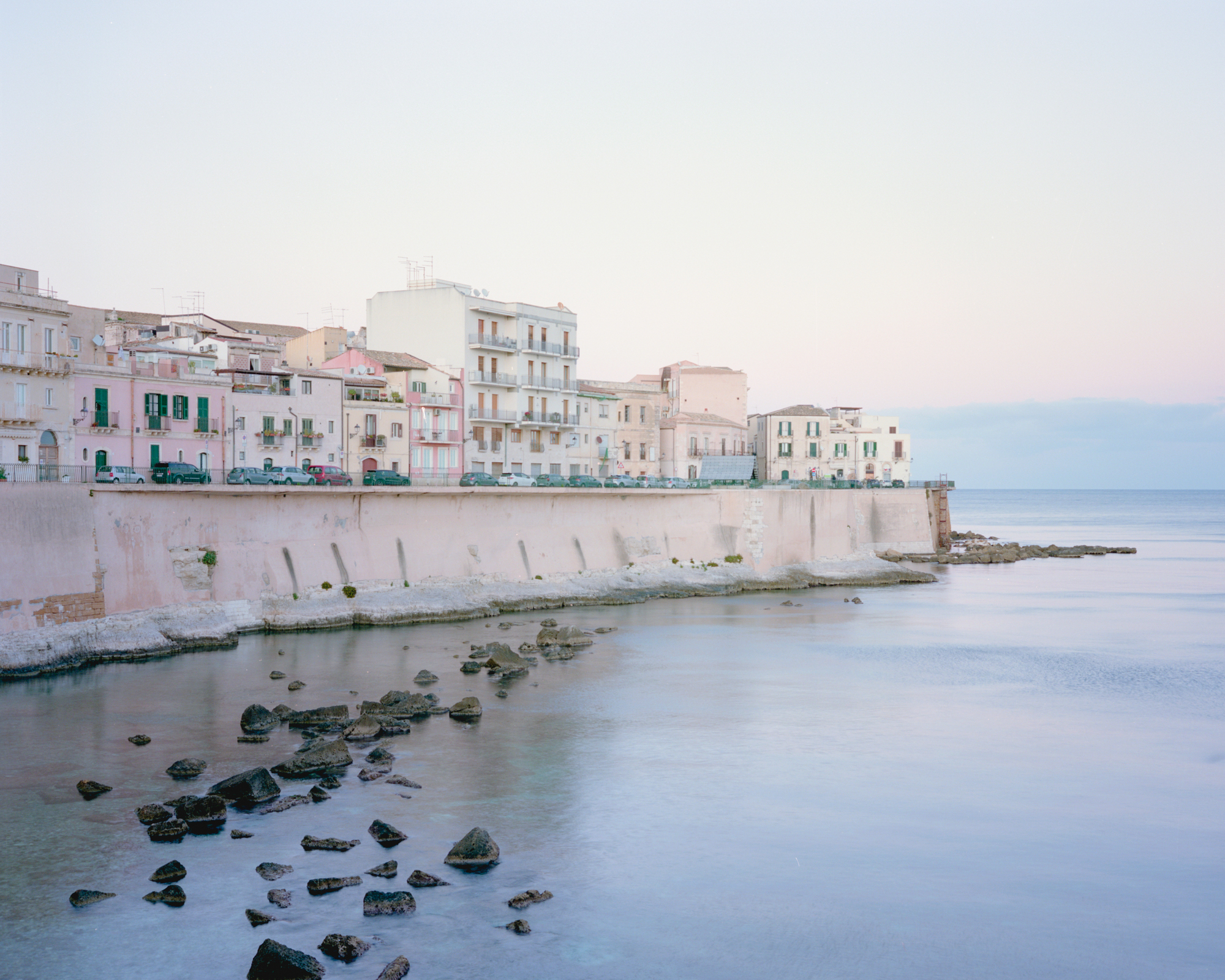 Waterfront residential buildings at dusk with exposed rocks in the shallows below the seawall