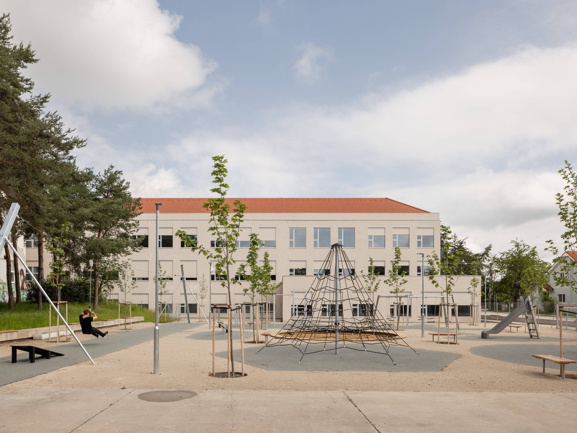 Playground with pyramid climbing structure and young trees facing a white plastered facade with terracotta roof