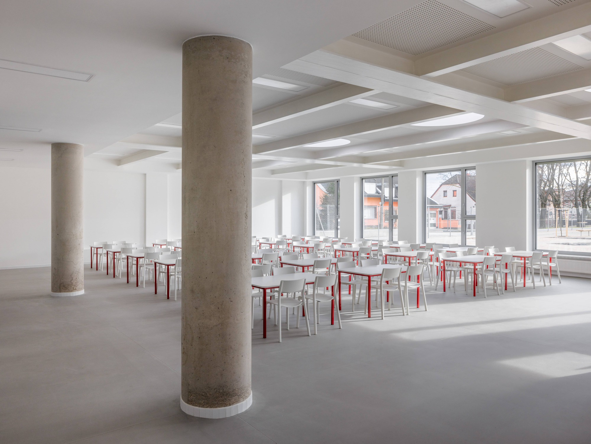 Interior dining hall with cylindrical concrete columns and rows of red and white chairs below a coffered ceiling