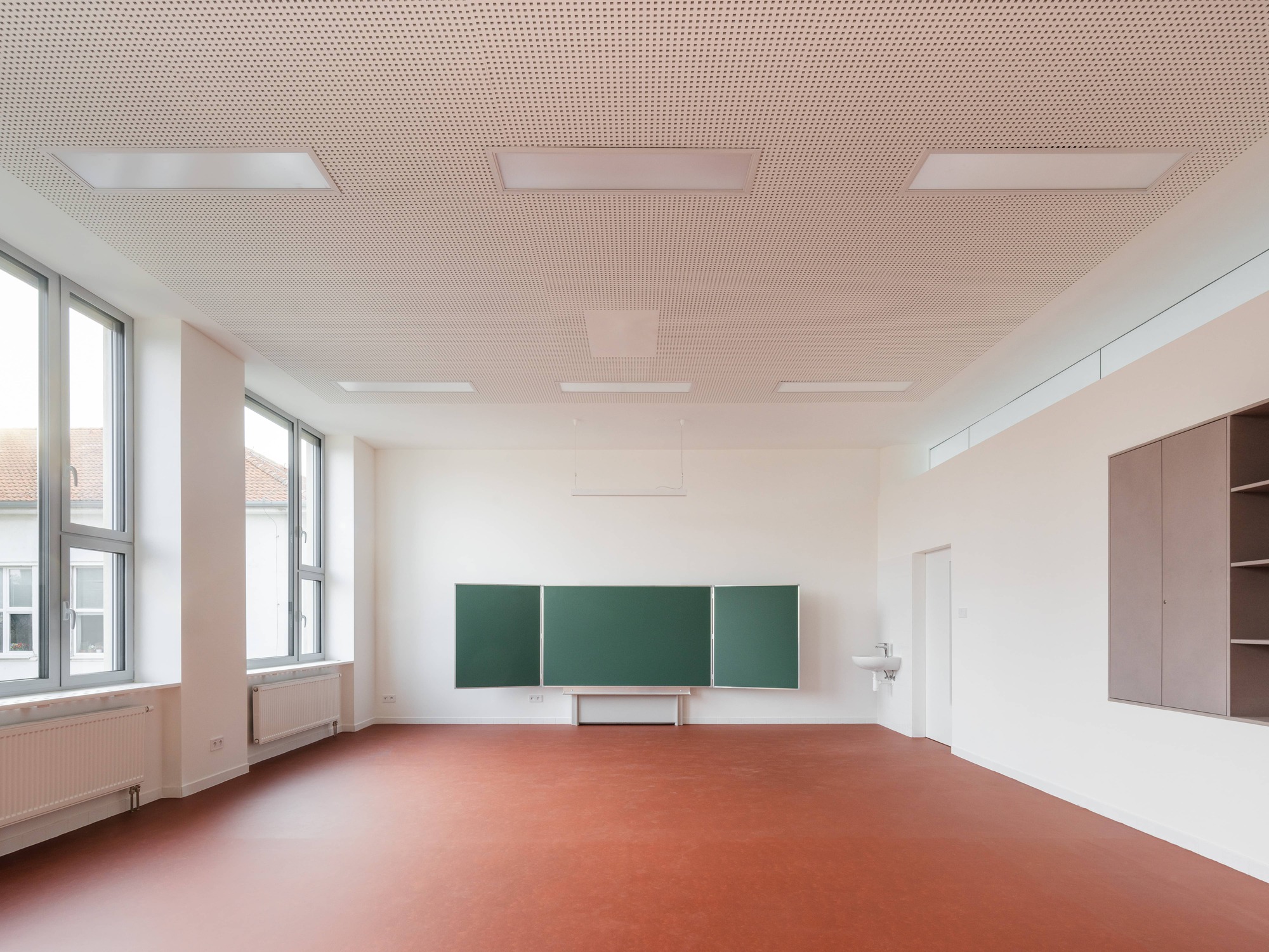 Classroom with green chalkboard panels mounted on white walls and terracotta linoleum flooring beneath a perforated acoustic ceiling
