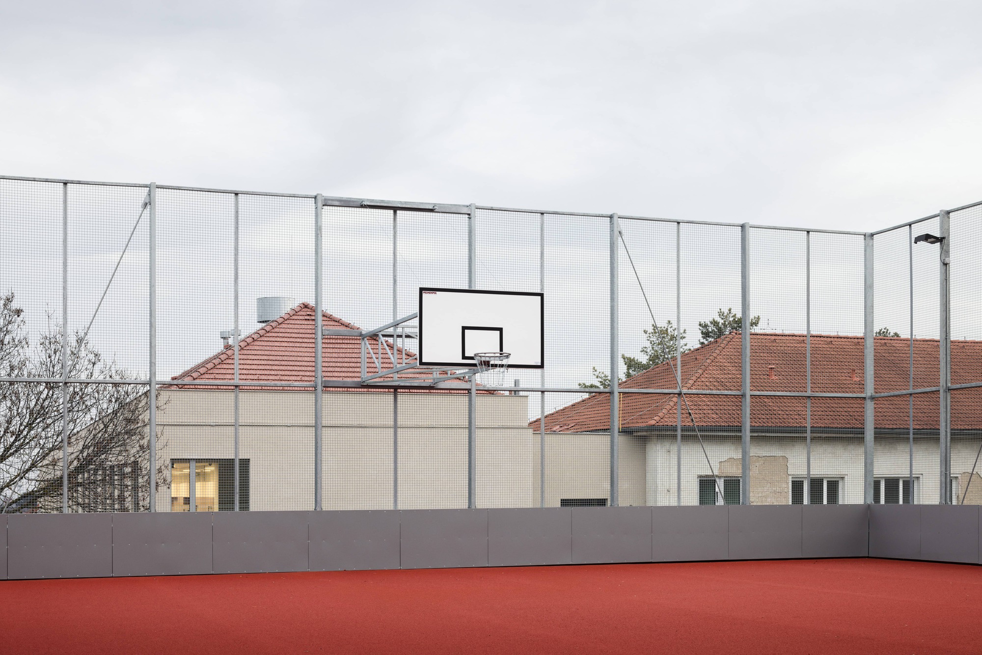 Rooftop sports court with red surface and basketball hoop behind wire mesh fencing under overcast sky