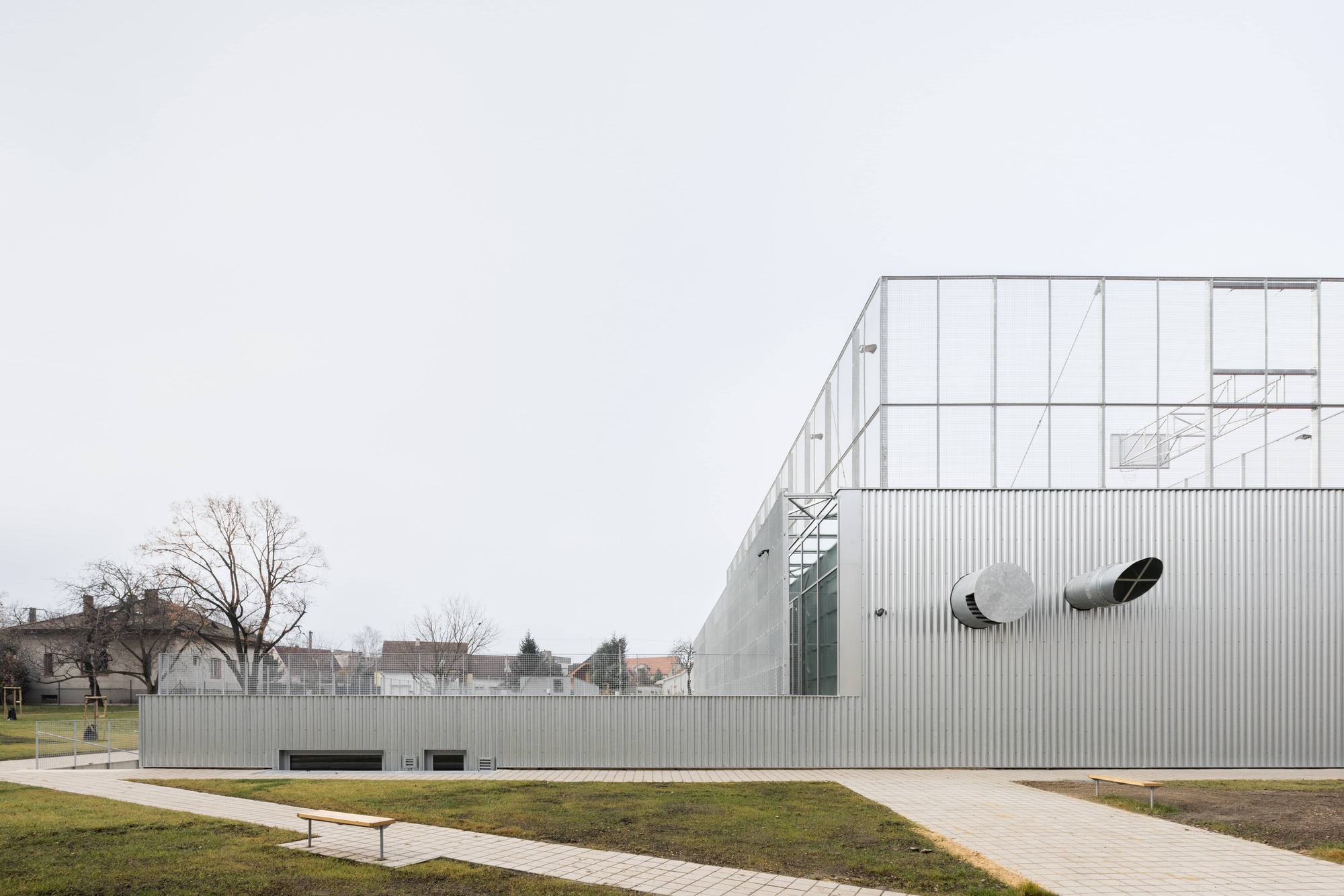 Corrugated metal facade with circular openings across a winter lawn with bare trees