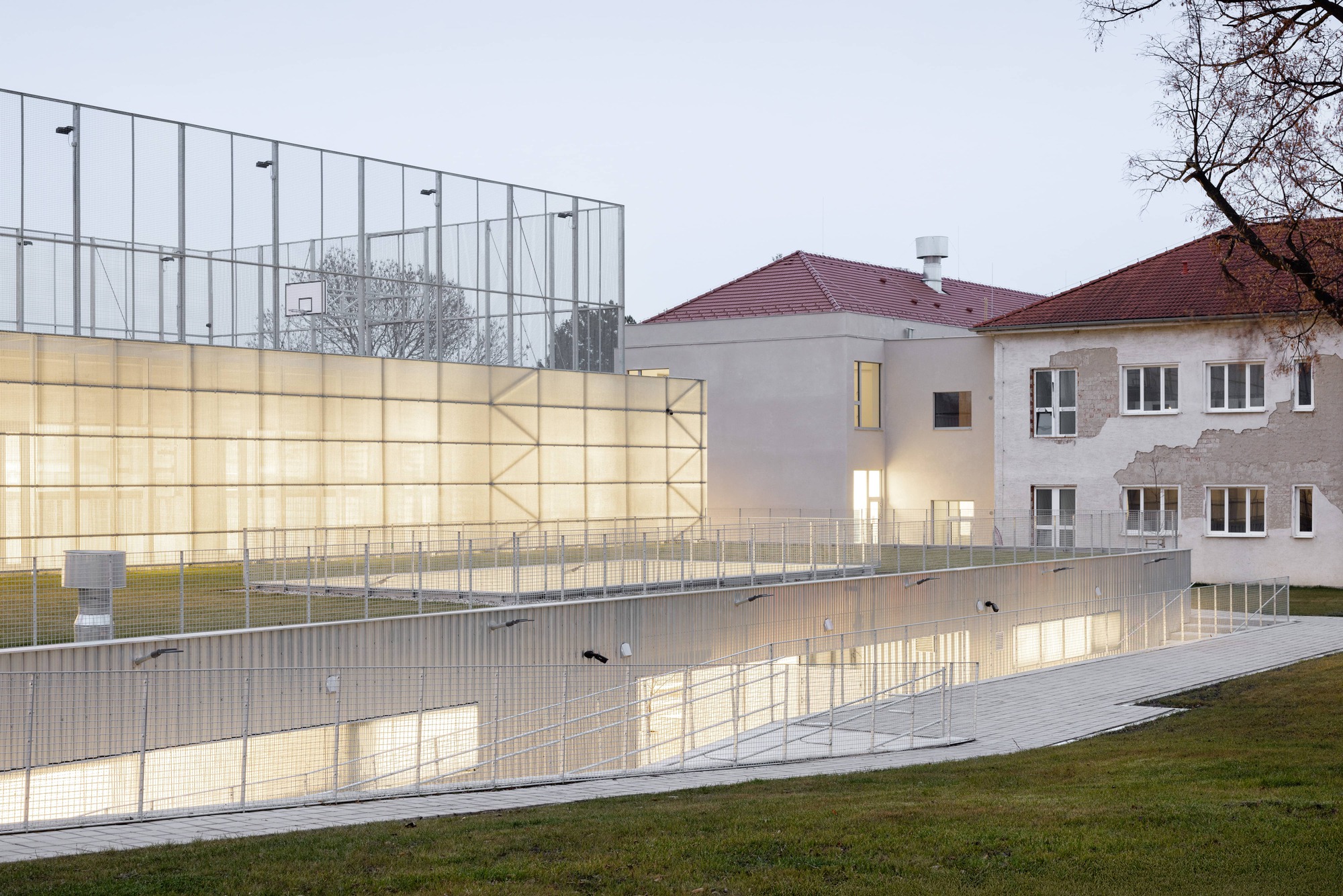 Translucent polycarbonate wall glowing at dusk beside an existing residential building with tiled roof