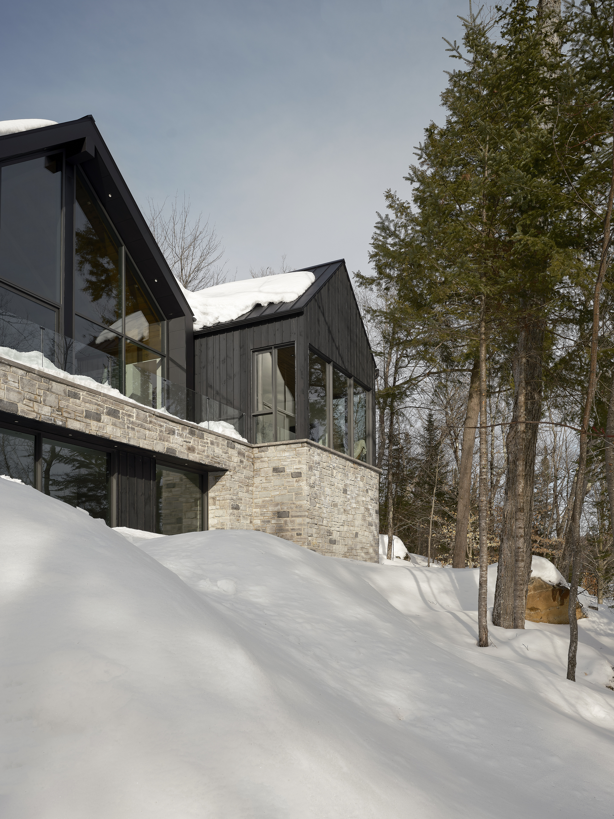 Stone base supporting dark clad upper volumes with pitched roofs on a snowy wooded slope