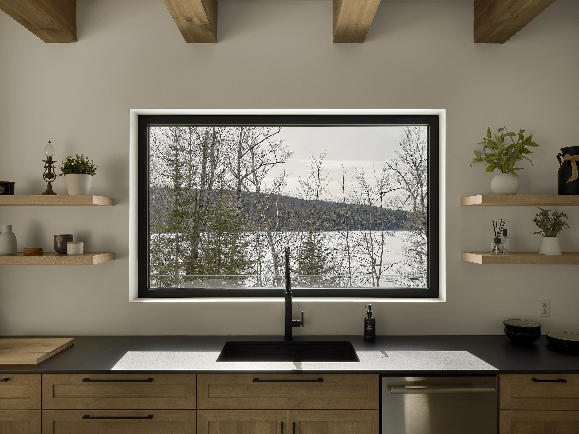Kitchen sink and black-framed window with floating timber shelves framing a view to winter lake