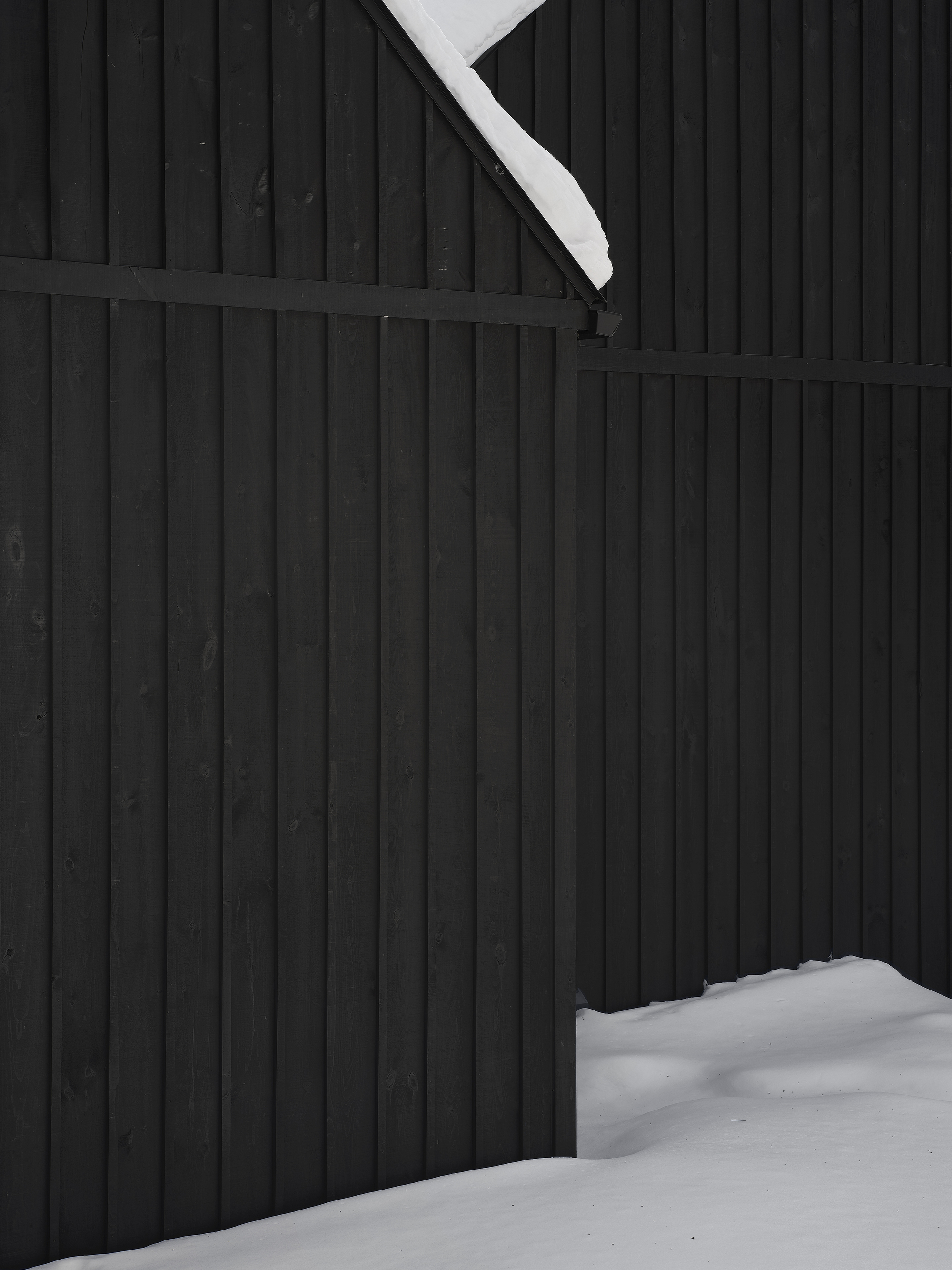 Close-up of black vertical wood siding meeting snow-covered roof edge and ground plane