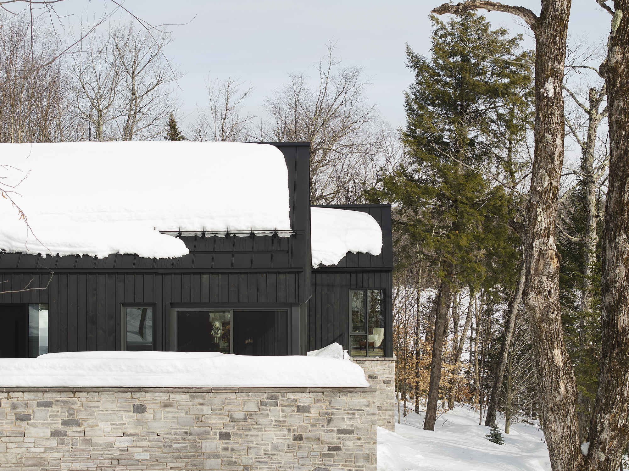 Stone base wall supporting black vertical wood cladding and snow-covered roof among bare trees