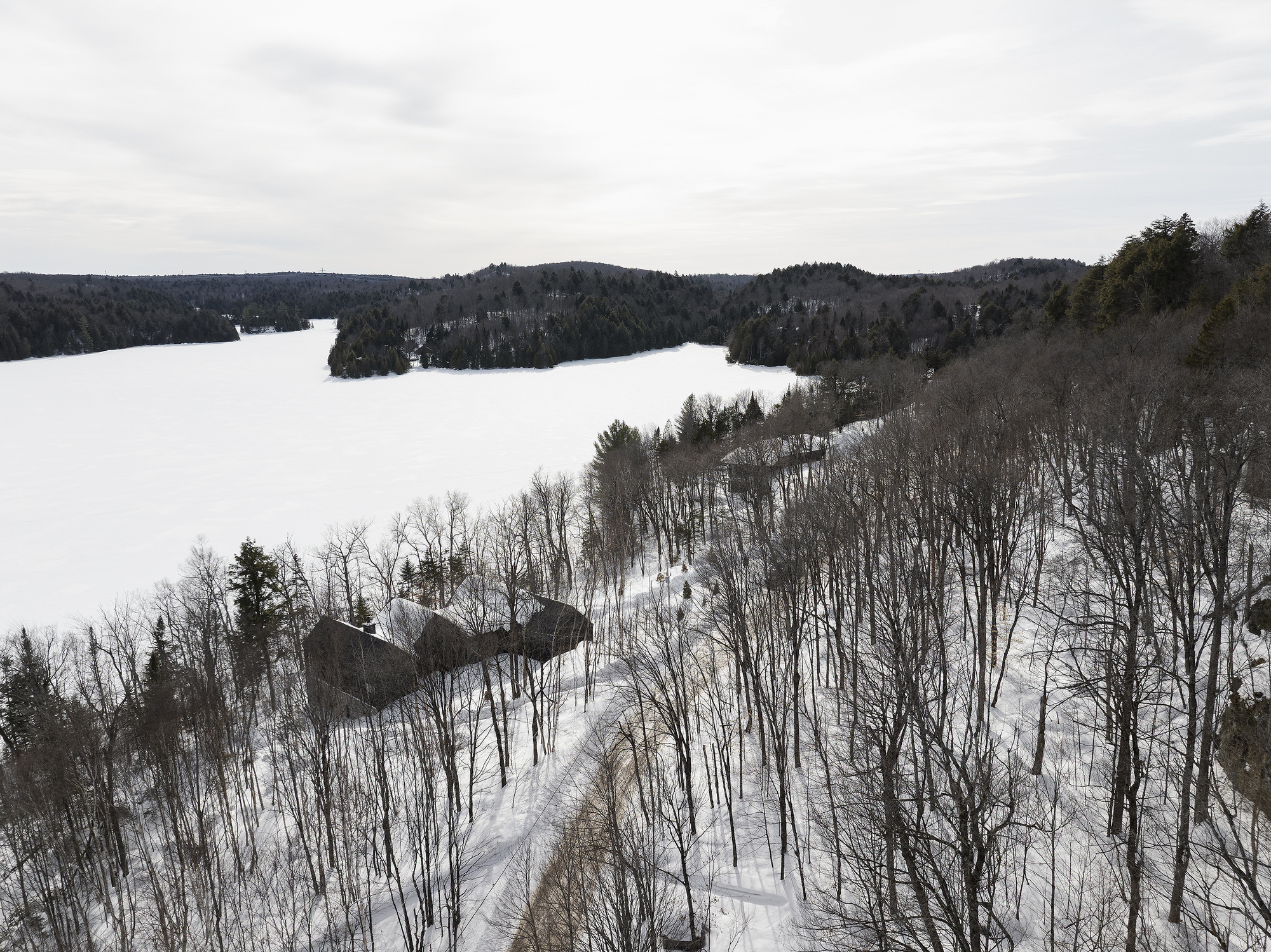 Aerial view of the dark pitched roof volumes nestled among bare winter trees beside a frozen lake