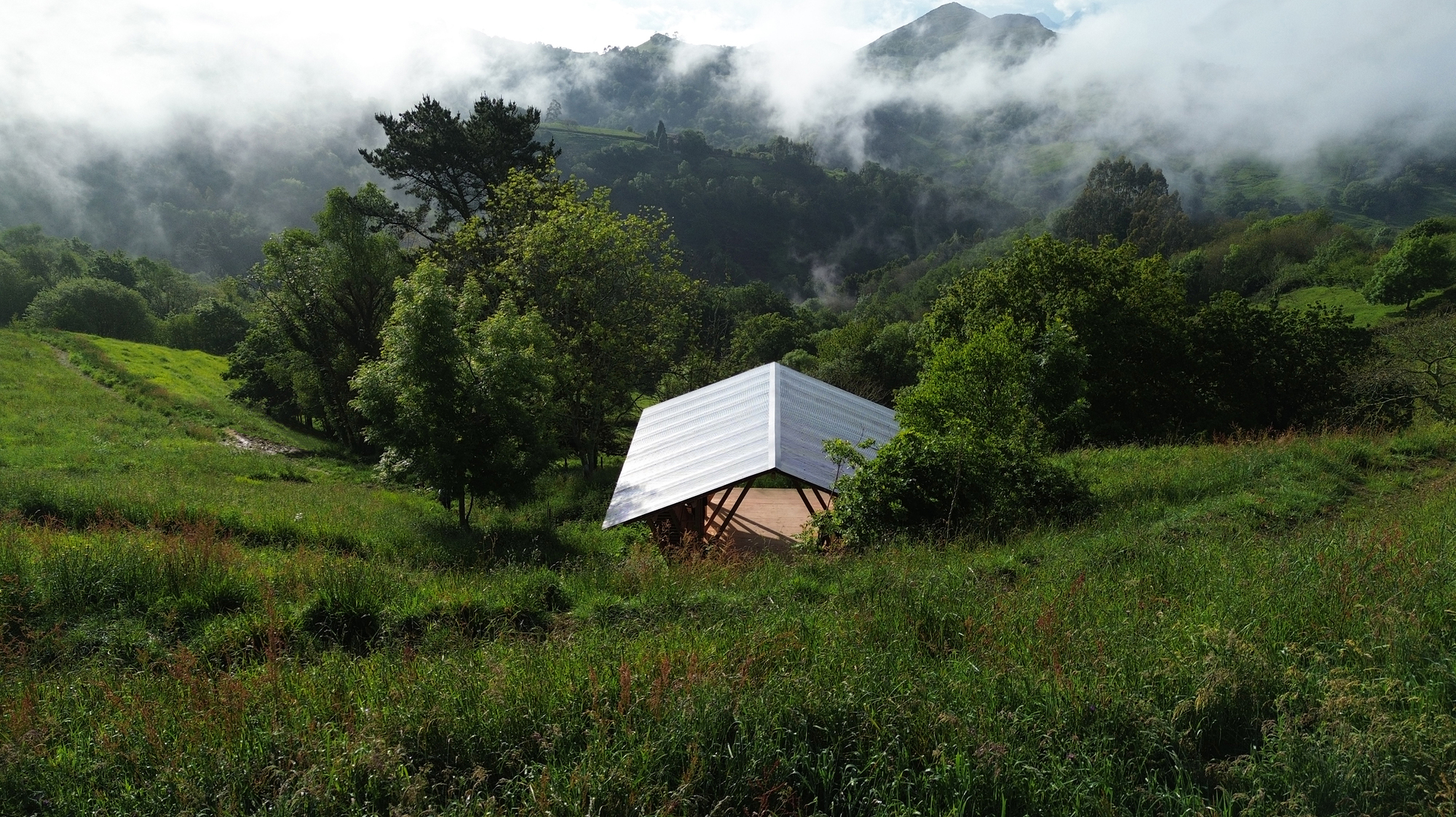 Small tilted pavilion with corrugated metal roof set into a green hillside with mist drifting through the trees