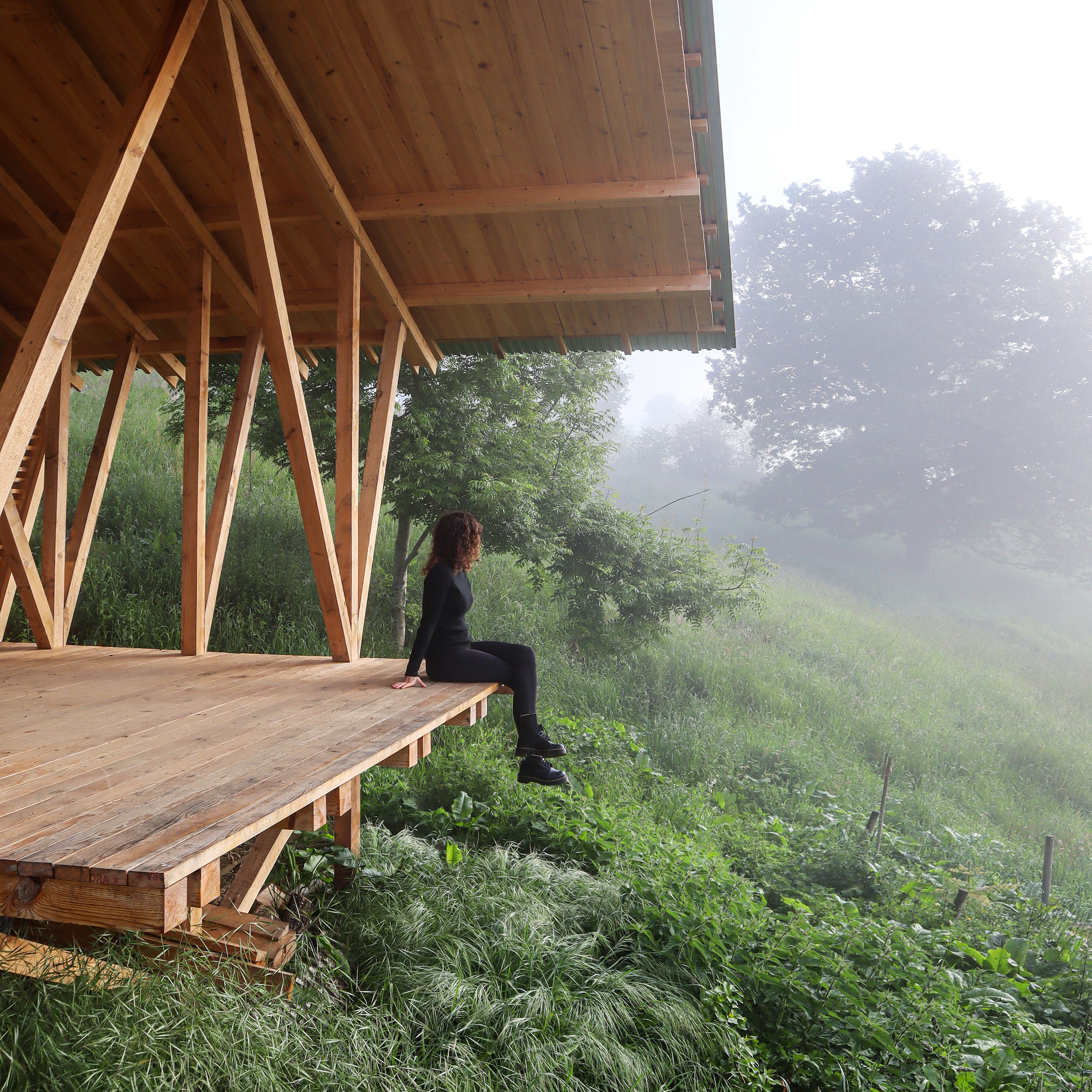 Person seated on the timber deck edge beneath the pavilion canopy in morning fog