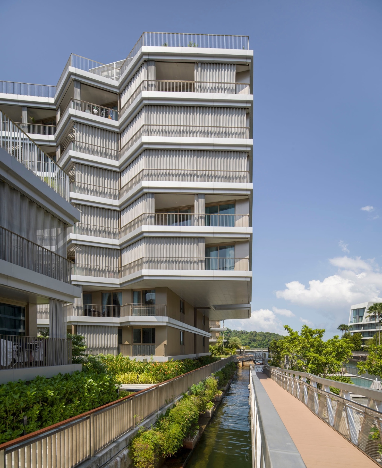 Zigzagging facade with vertical louvers and recessed balconies above a planted canal under blue sky