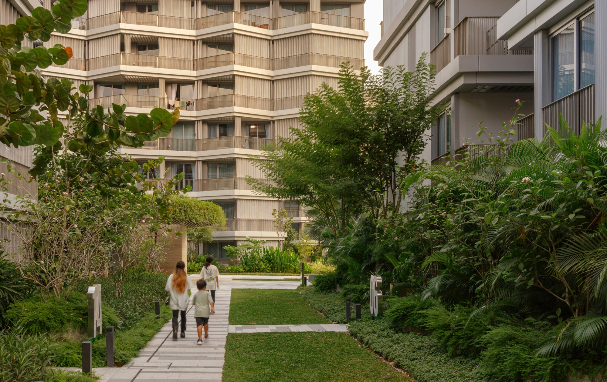 Pathway through a planted courtyard with two residents walking past residential towers under soft daylight