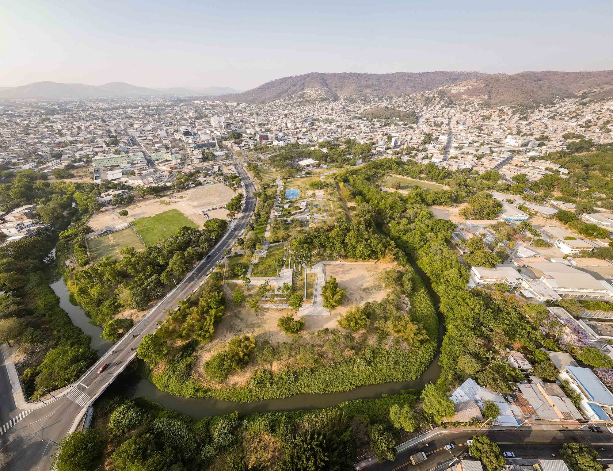 Aerial view of river-bounded park with dense tree cover cutting through adjacent urban development and hills
