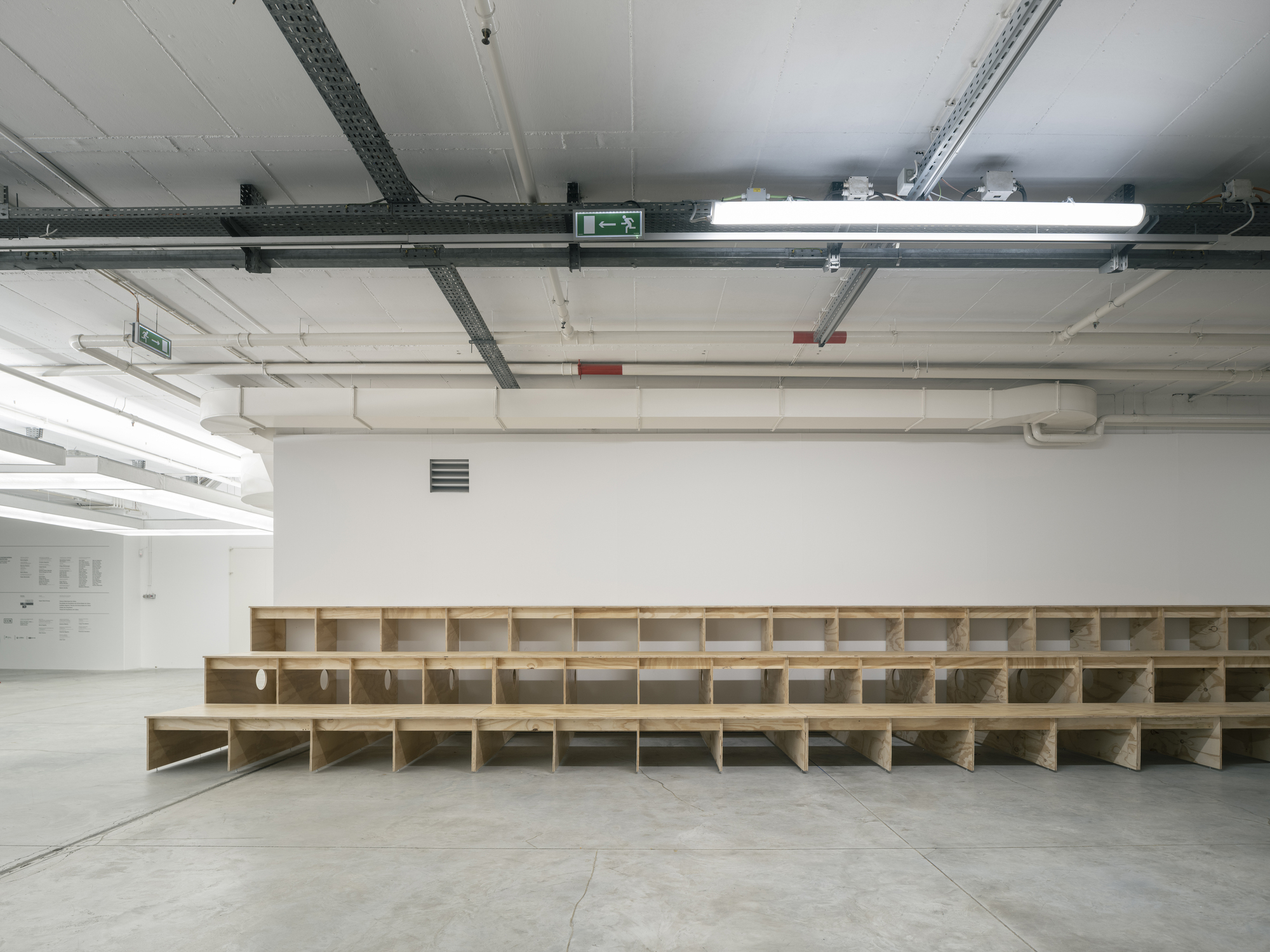 Long tiered plywood seating structure with stepped risers spanning the width of a white gallery space