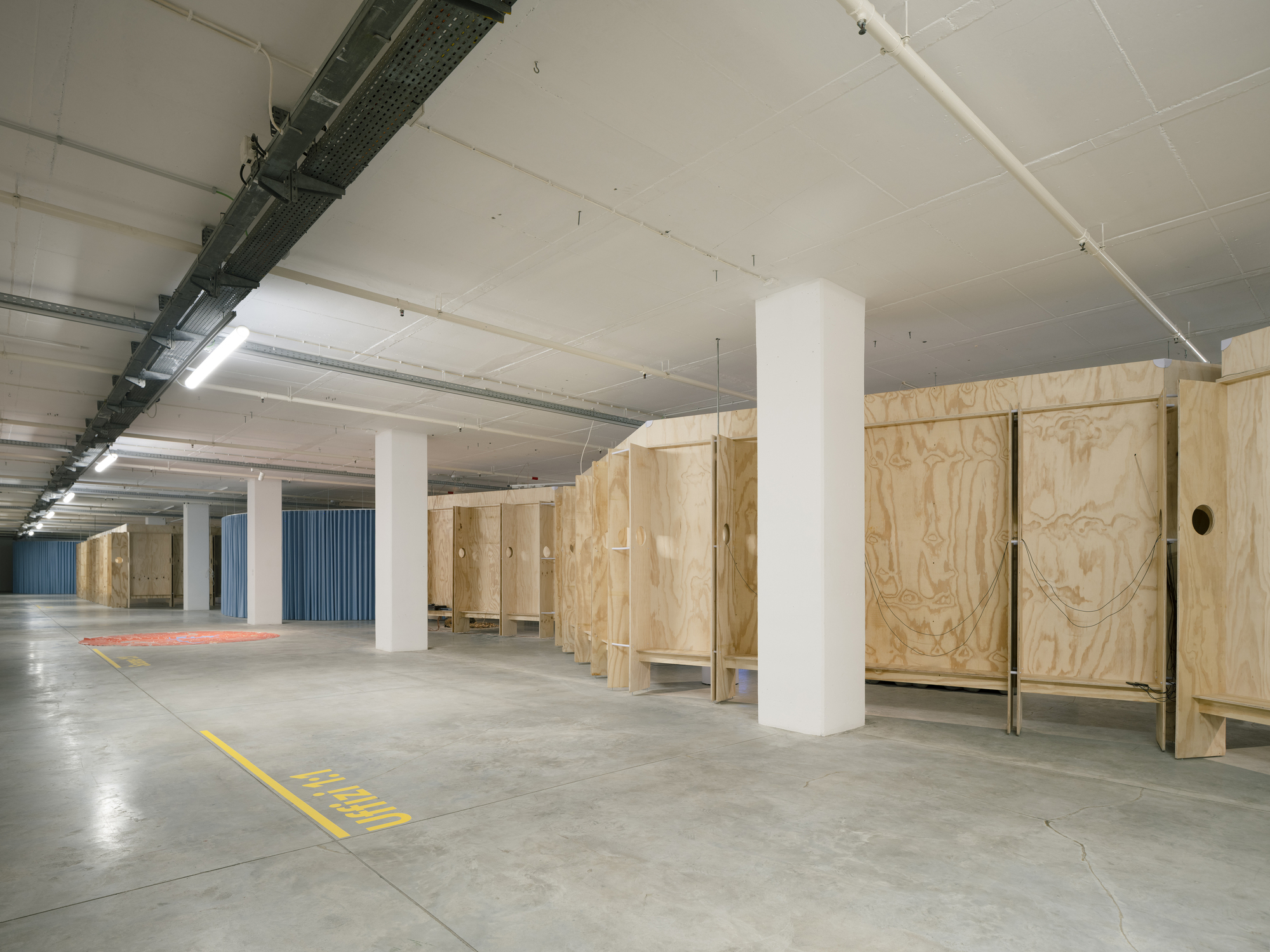Open gallery space with white cylindrical columns and freestanding plywood storage units under fluorescent lighting