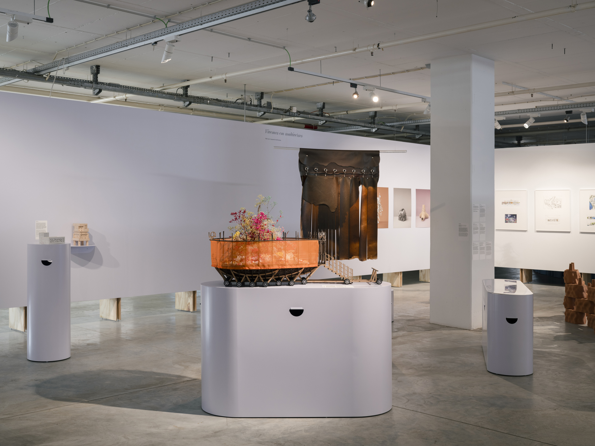 Exhibition space with sculptural installation on white plinth beneath exposed ceiling ducts and track lights