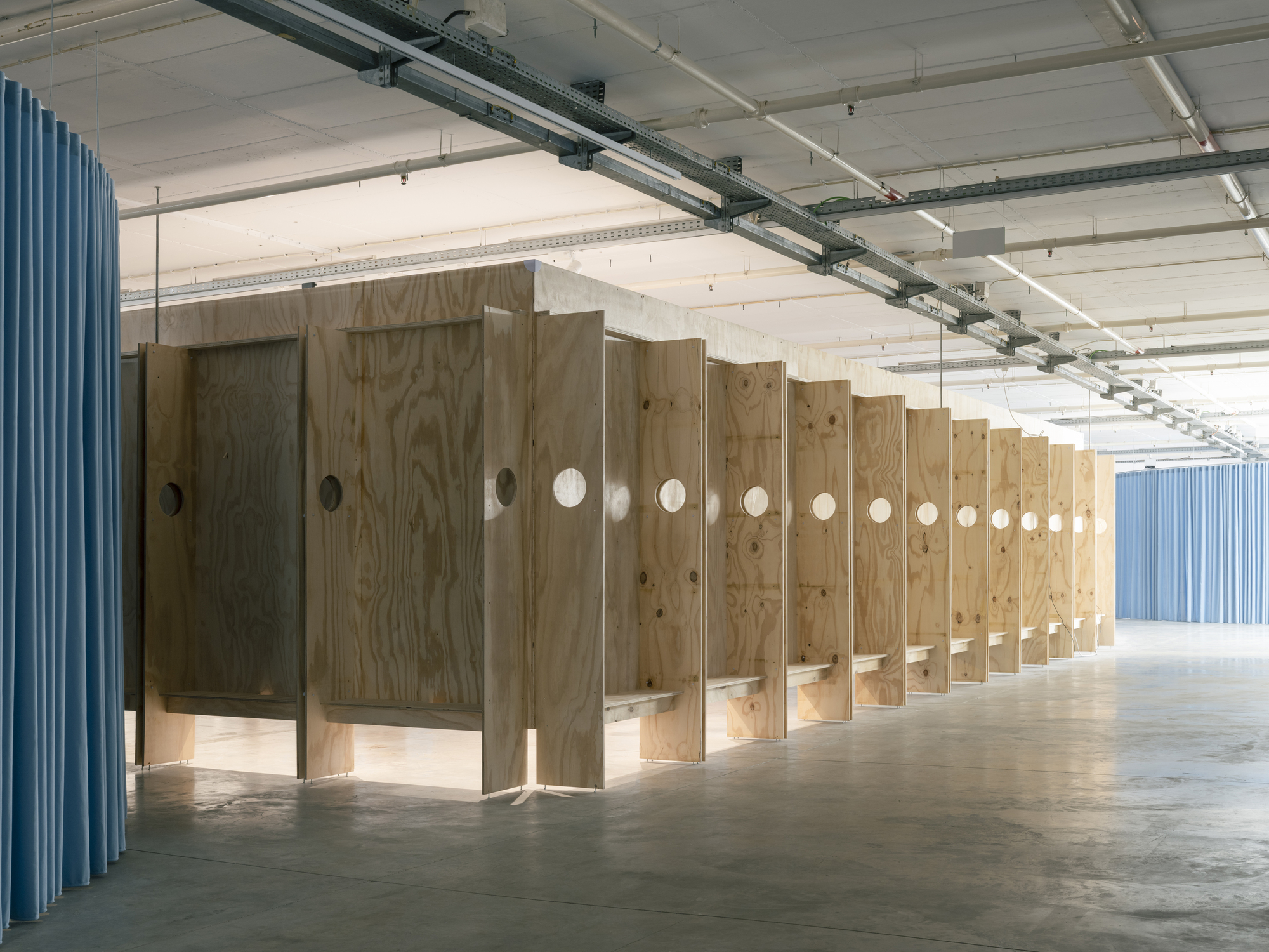 Row of plywood partitions with circular openings flanked by blue corrugated walls under exposed concrete ceiling