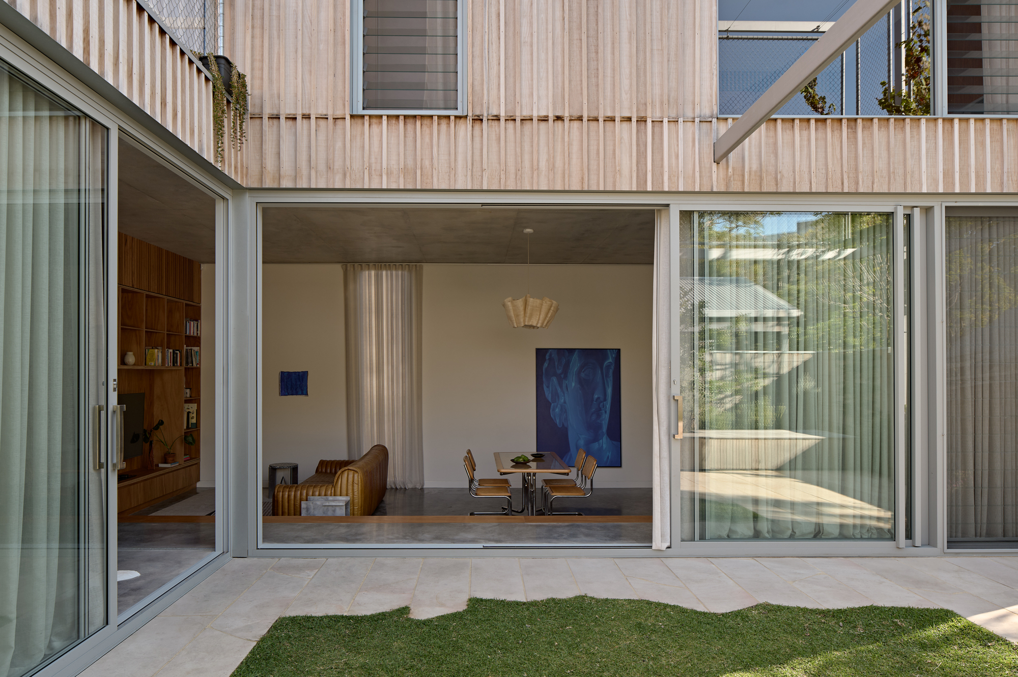 Sliding glass doors opening the interior to a paved courtyard with lawn and timber-clad upper volumes beyond