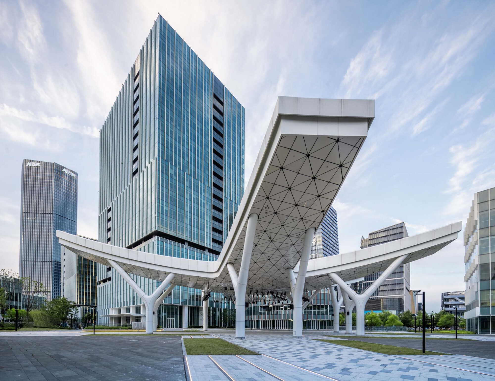 White triangulated canopy with branching columns sheltering the plaza beneath glass tower blocks under wispy clouds