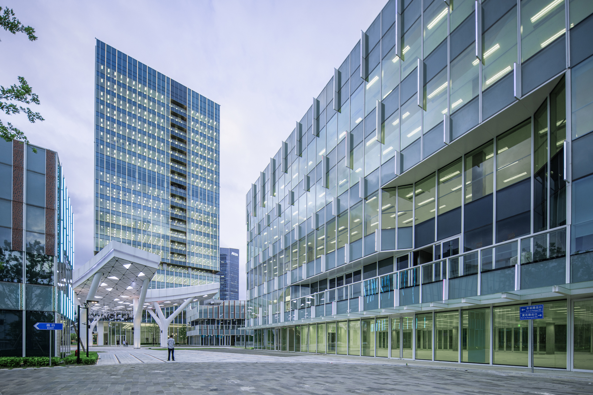 Wide plaza view with curtain wall towers and a white canopy structure supported by angled columns at dusk