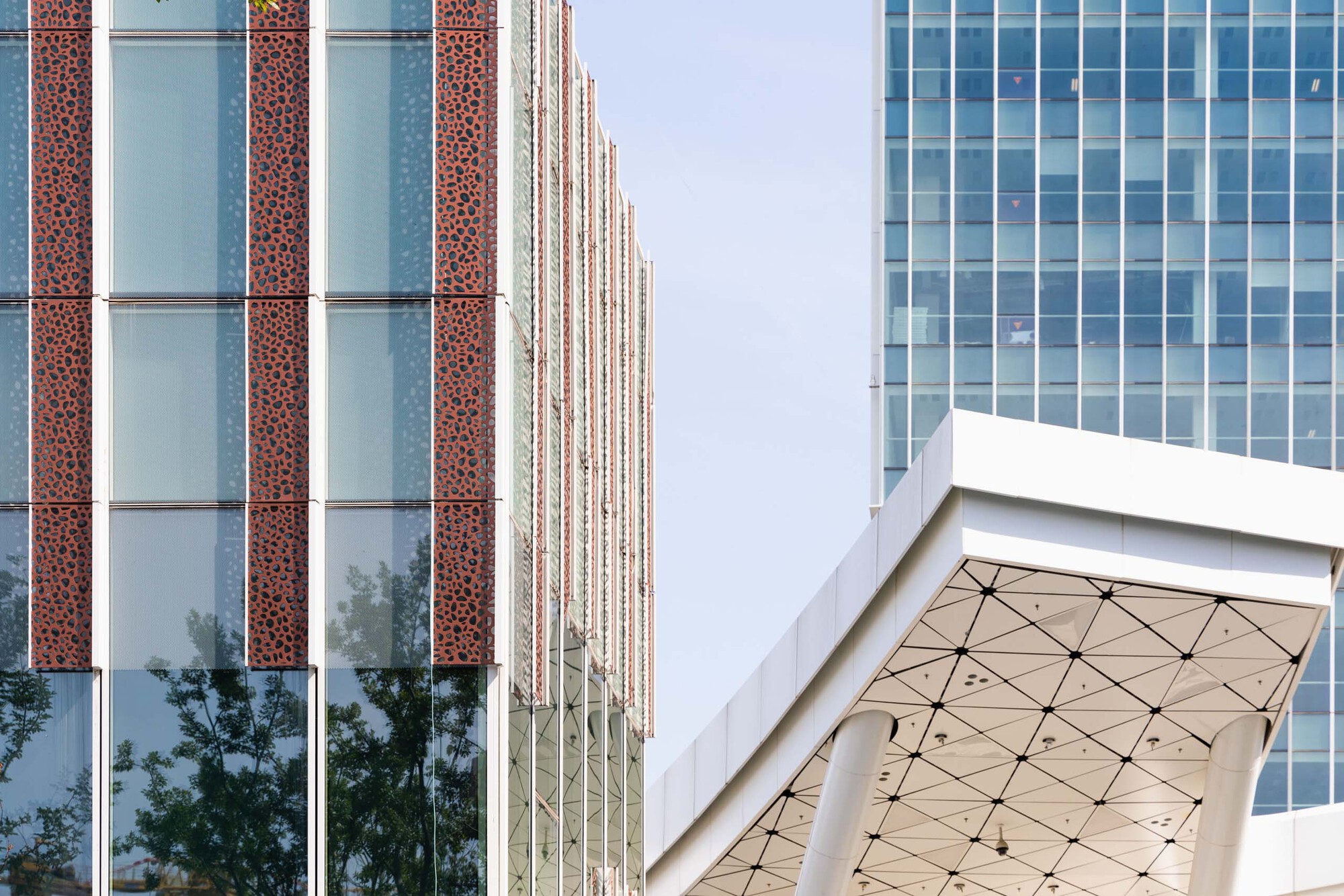 Facade detail showing perforated red panels alternating with glass curtain wall and triangulated canopy soffit