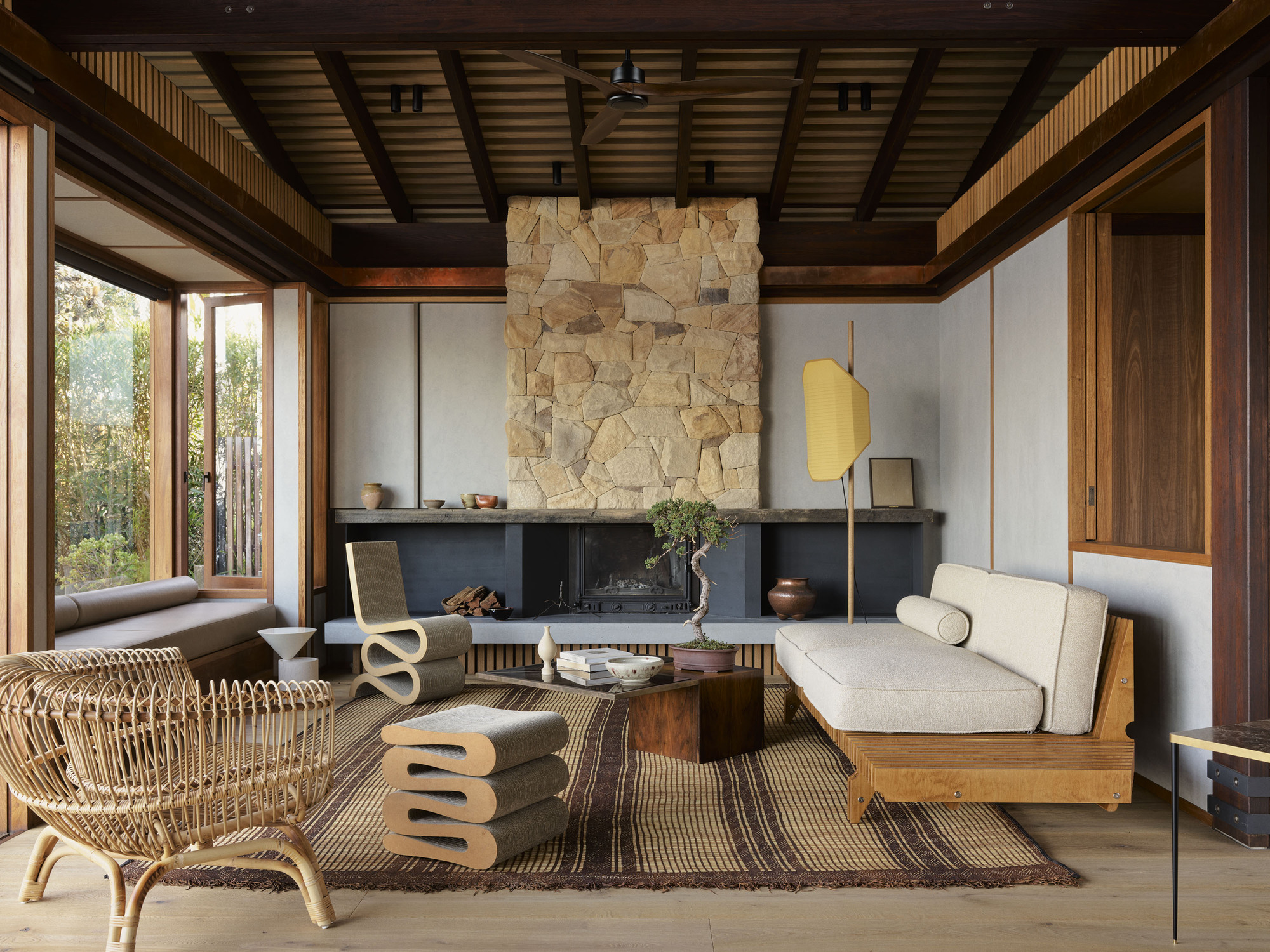 Living room with exposed timber ceiling beams and rough stone fireplace surround facing floor-to-ceiling windows