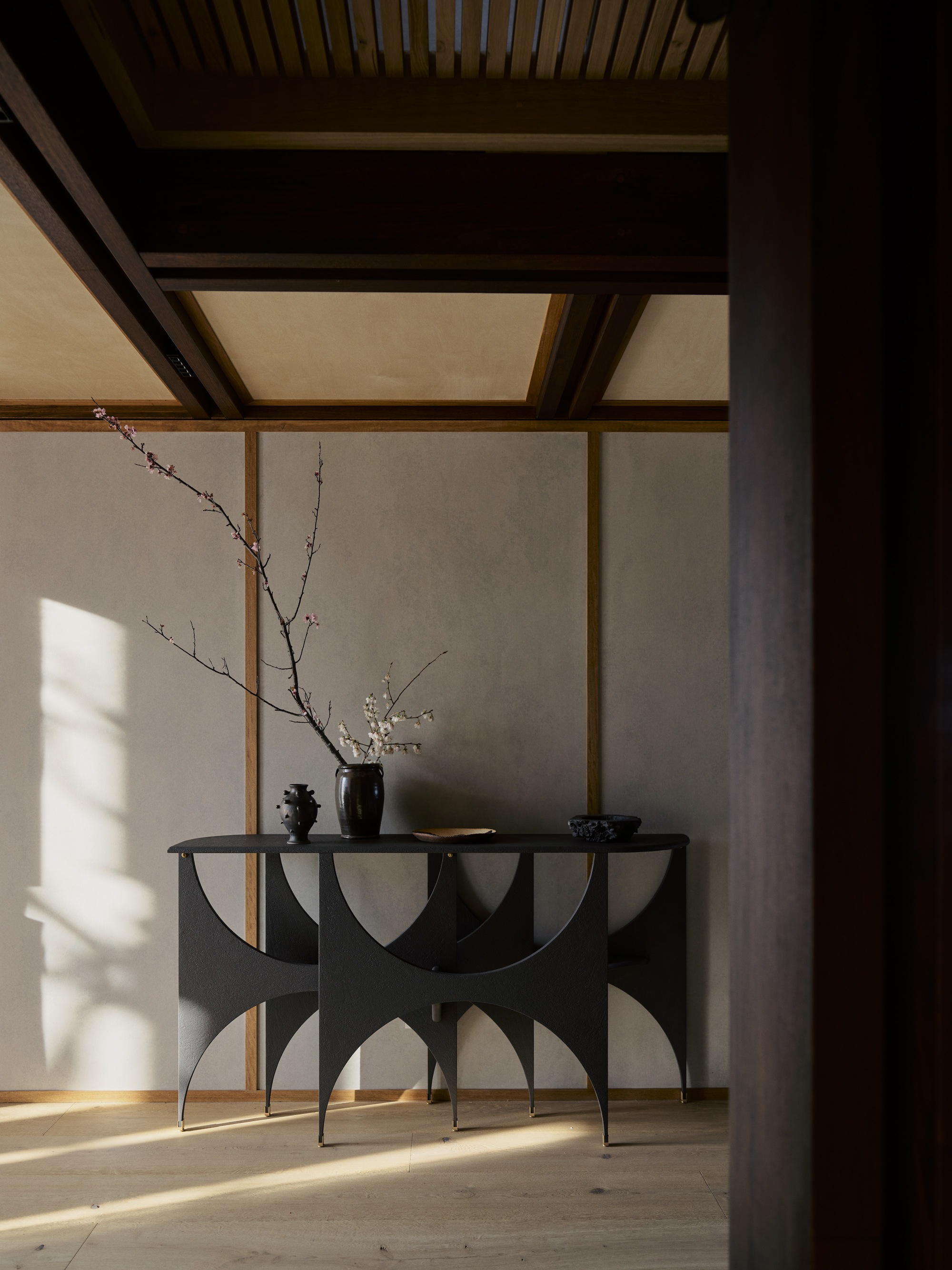 Sculptural black console table with flowering branch against timber-framed wall in morning light