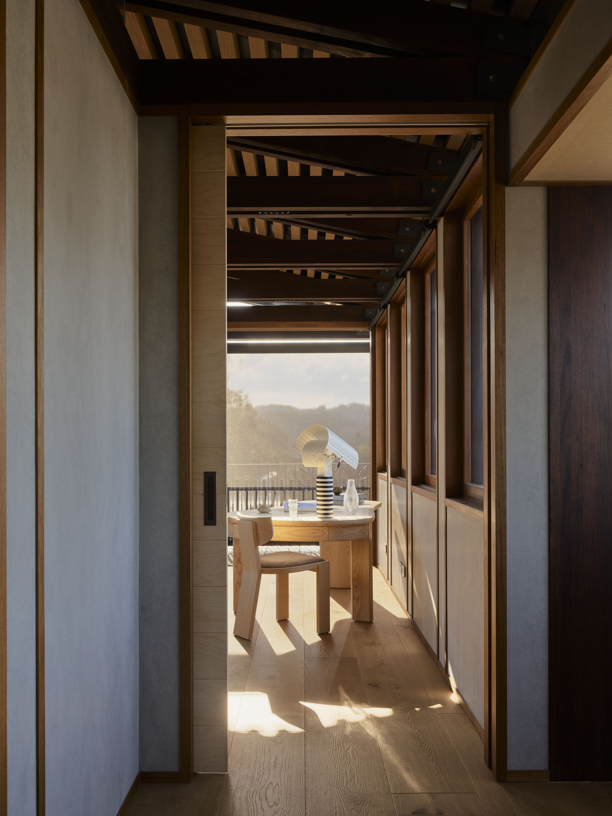 Narrow hallway with exposed timber ceiling beams leading to a desk and chair framed by sunlight