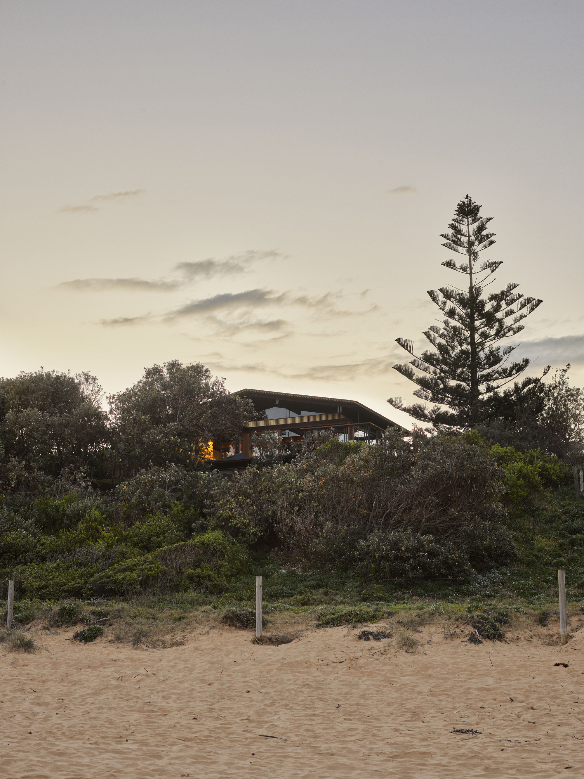 View from the beach showing the curved roof structure nestled among coastal vegetation at dusk