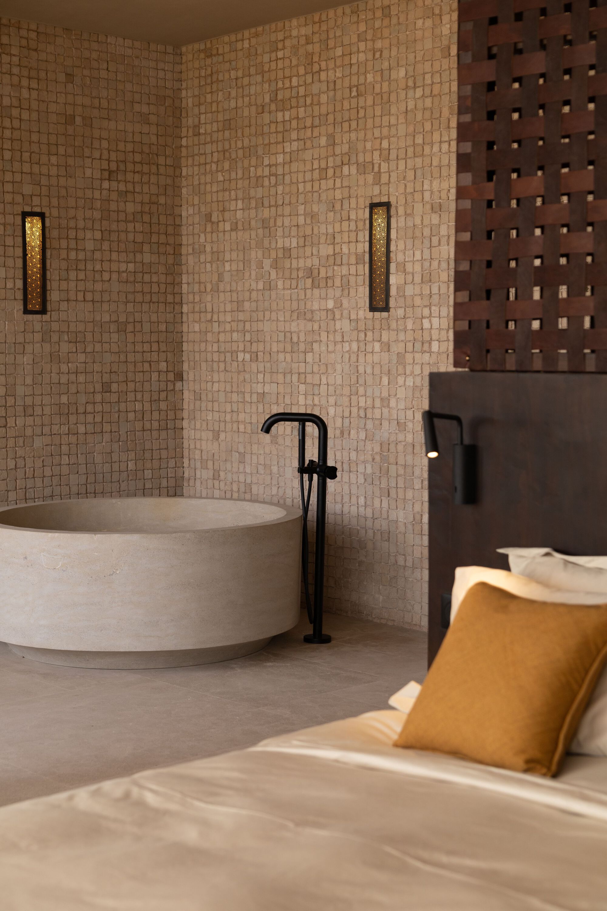 Circular stone bathtub with freestanding black tap against a small-tile mosaic wall in low light