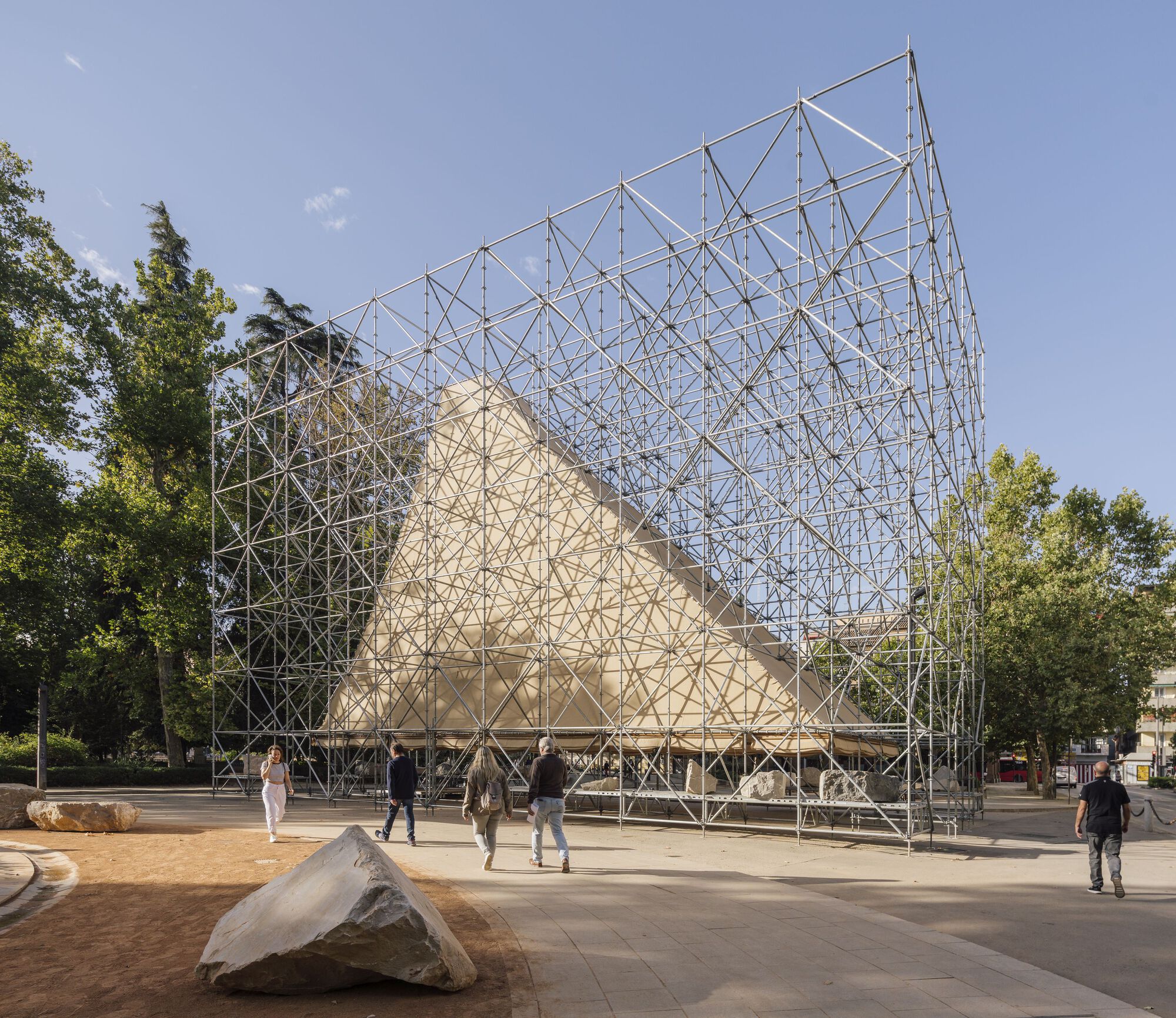 Triangular timber pavilion enclosed by a steel scaffolding framework in a public plaza with scattered stones