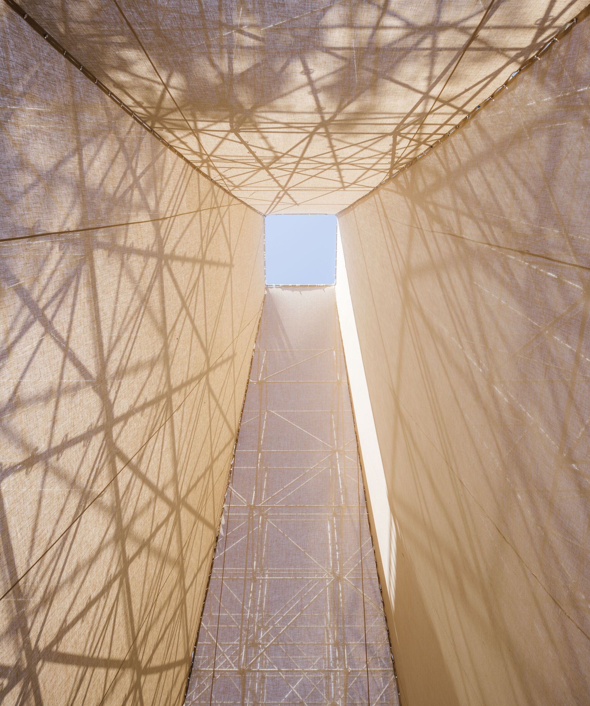 Upward view through the central void showing steel trusses and scaffold shadows on translucent fabric surfaces