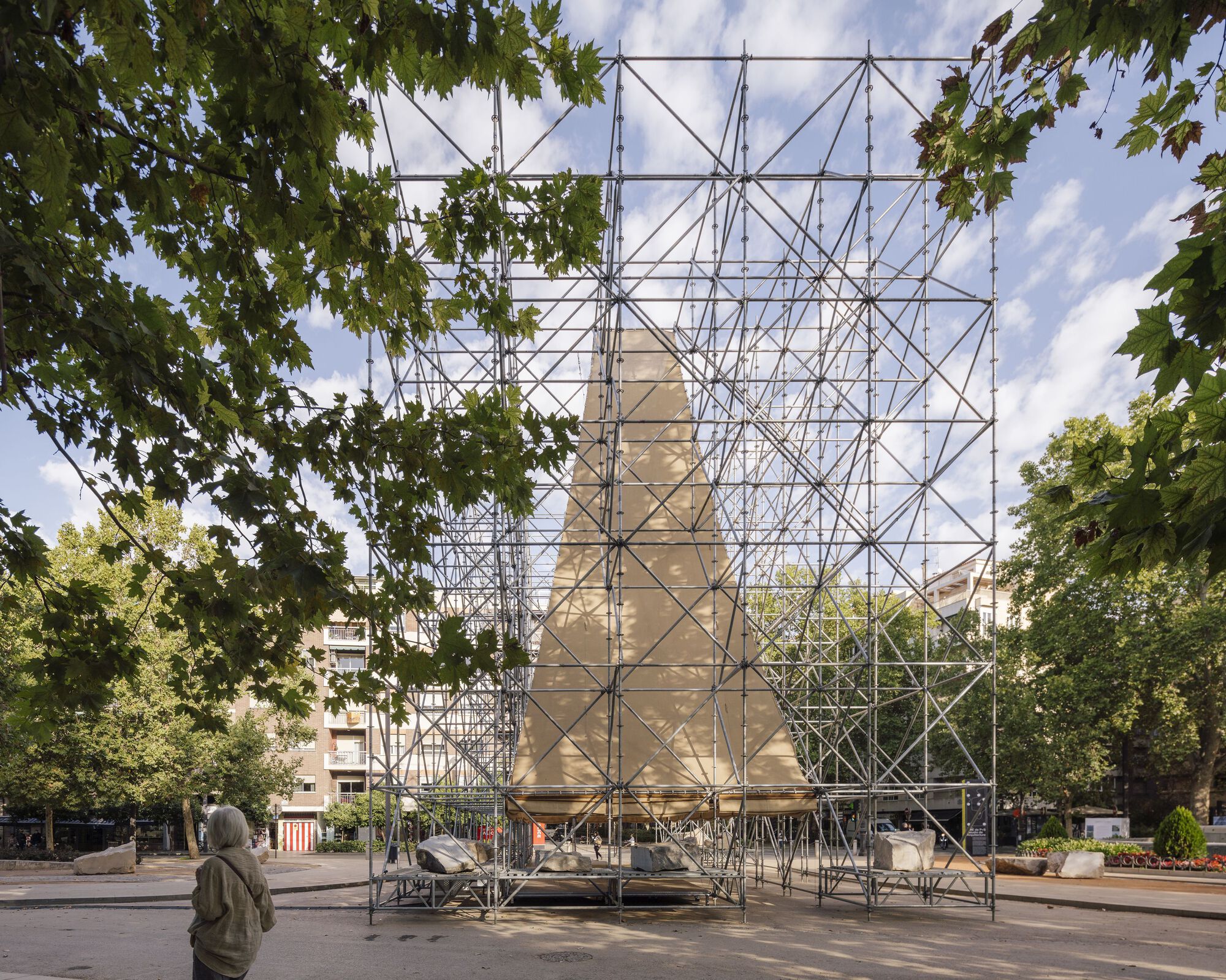 Steel tube scaffolding enclosing a pitched-roof form seen through overhanging tree branches with a visitor nearby