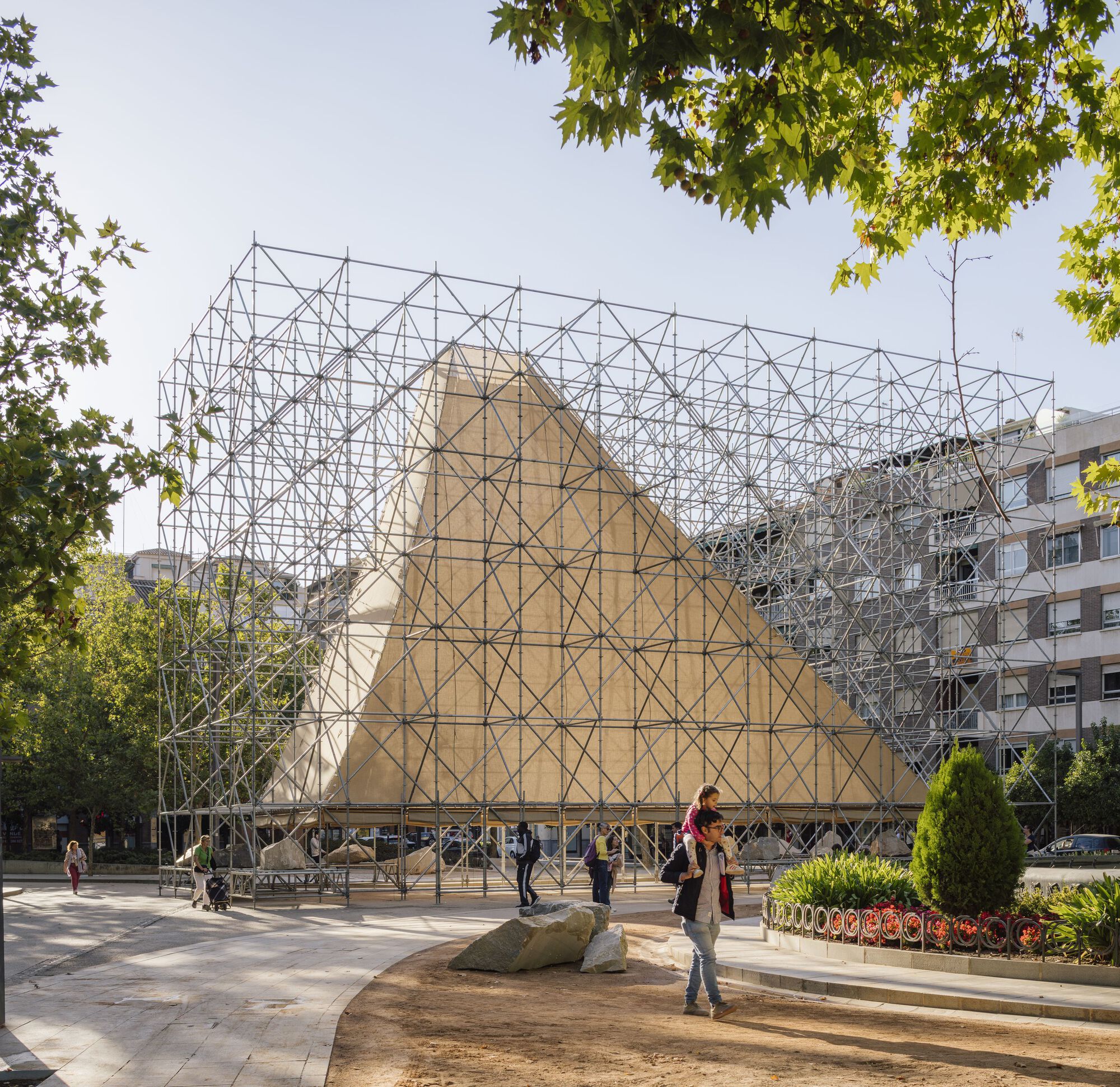 Triangulated metal scaffold pavilion with plywood-clad gable in a plaza with large boulders and planted beds