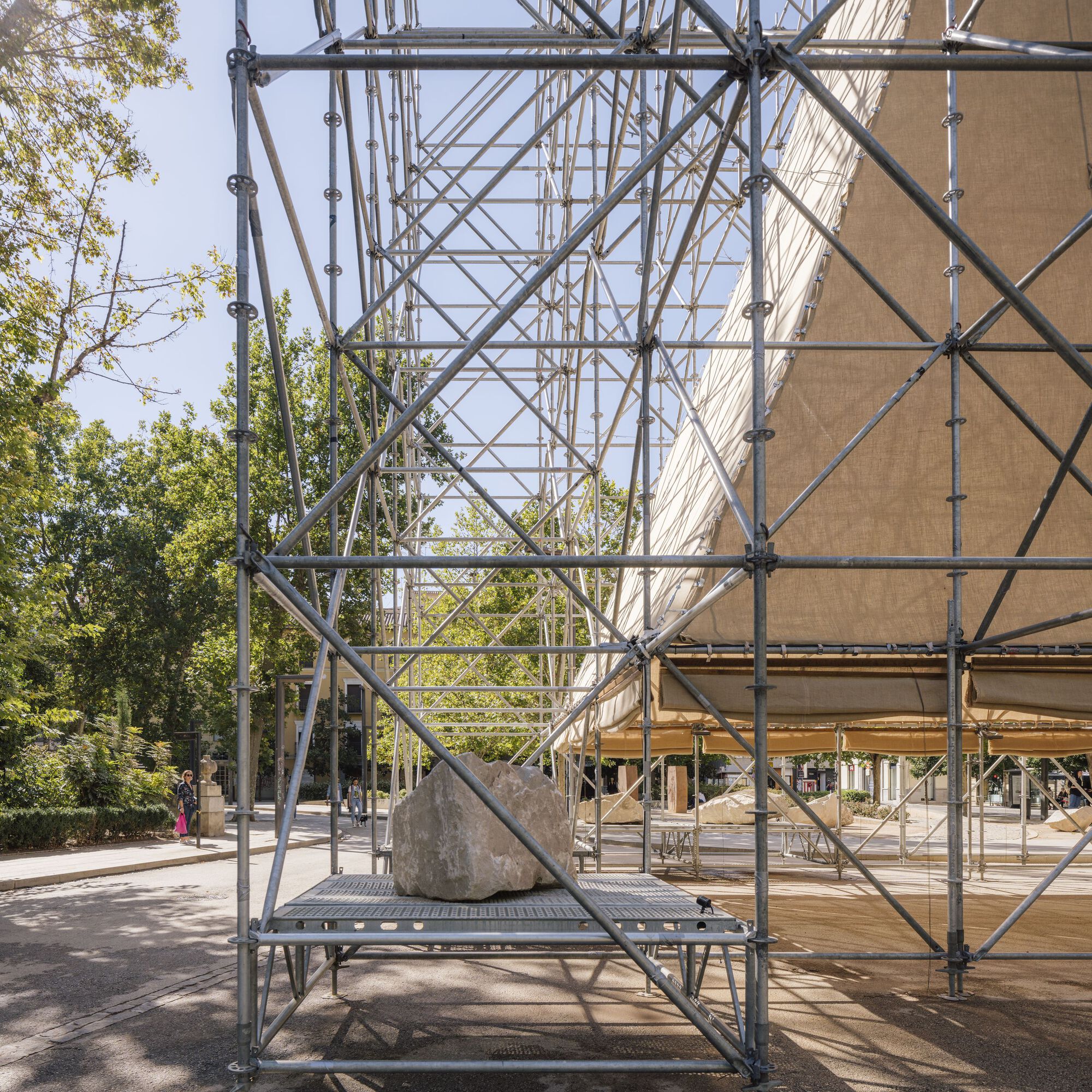 Metal scaffold frame detail with large boulder on platform and tree foliage visible beyond