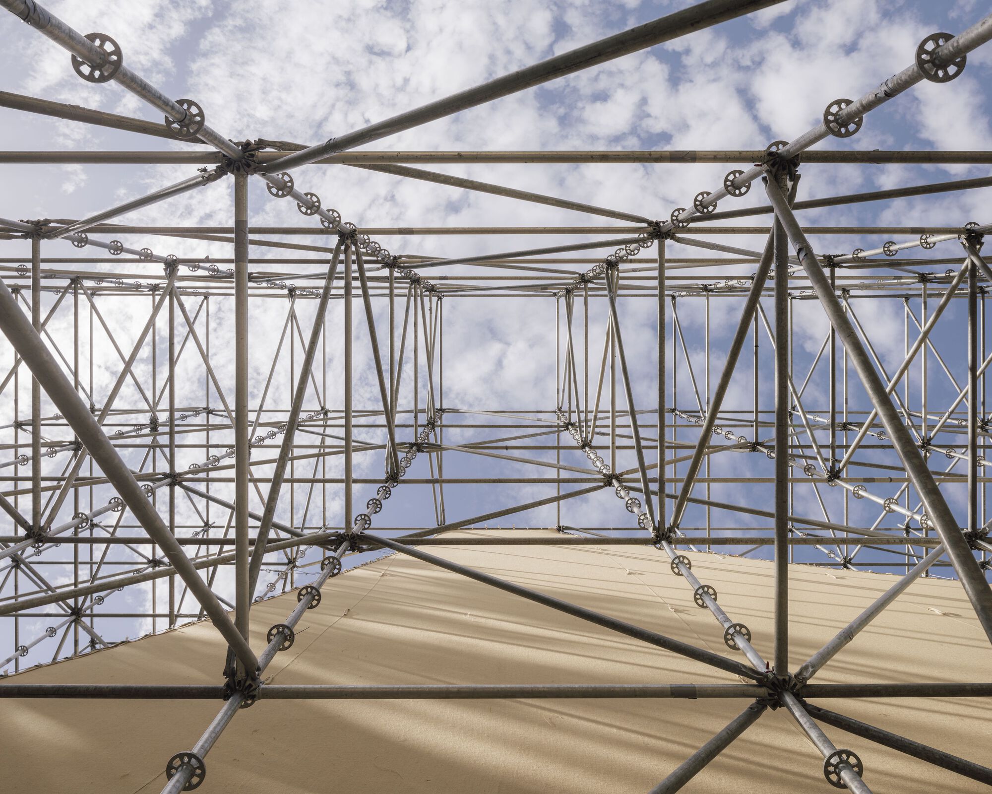 Upward view into scaffolding grid with circular connector nodes and plywood panels against blue sky
