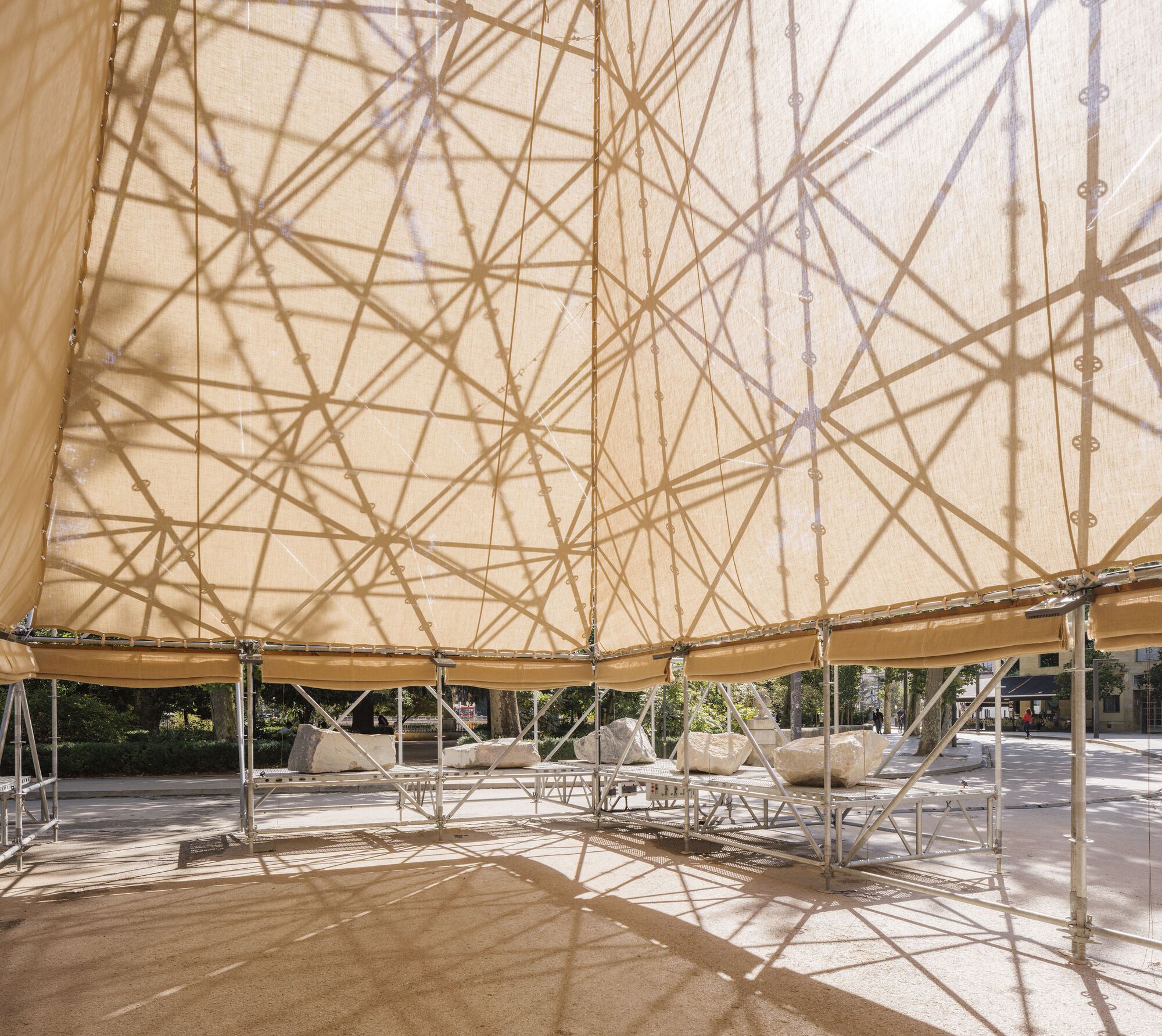 Dappled light patterns from the fabric roof overhead illuminating hammocks suspended beneath the pavilion structure