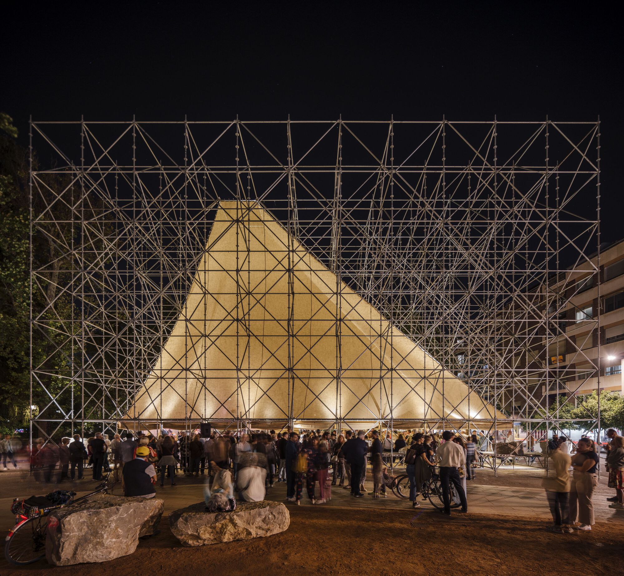 Nighttime view of the glowing scaffold-clad pavilion with crowds gathered around its base
