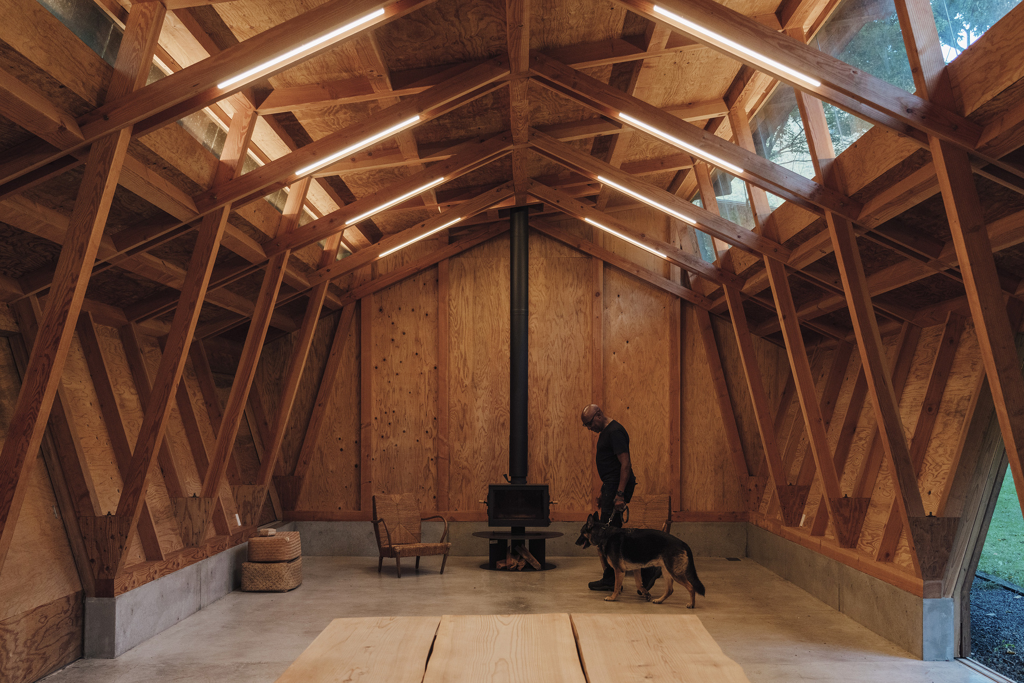 Main hall with a person and dog beside a central stove under the pitched timber frame at dusk