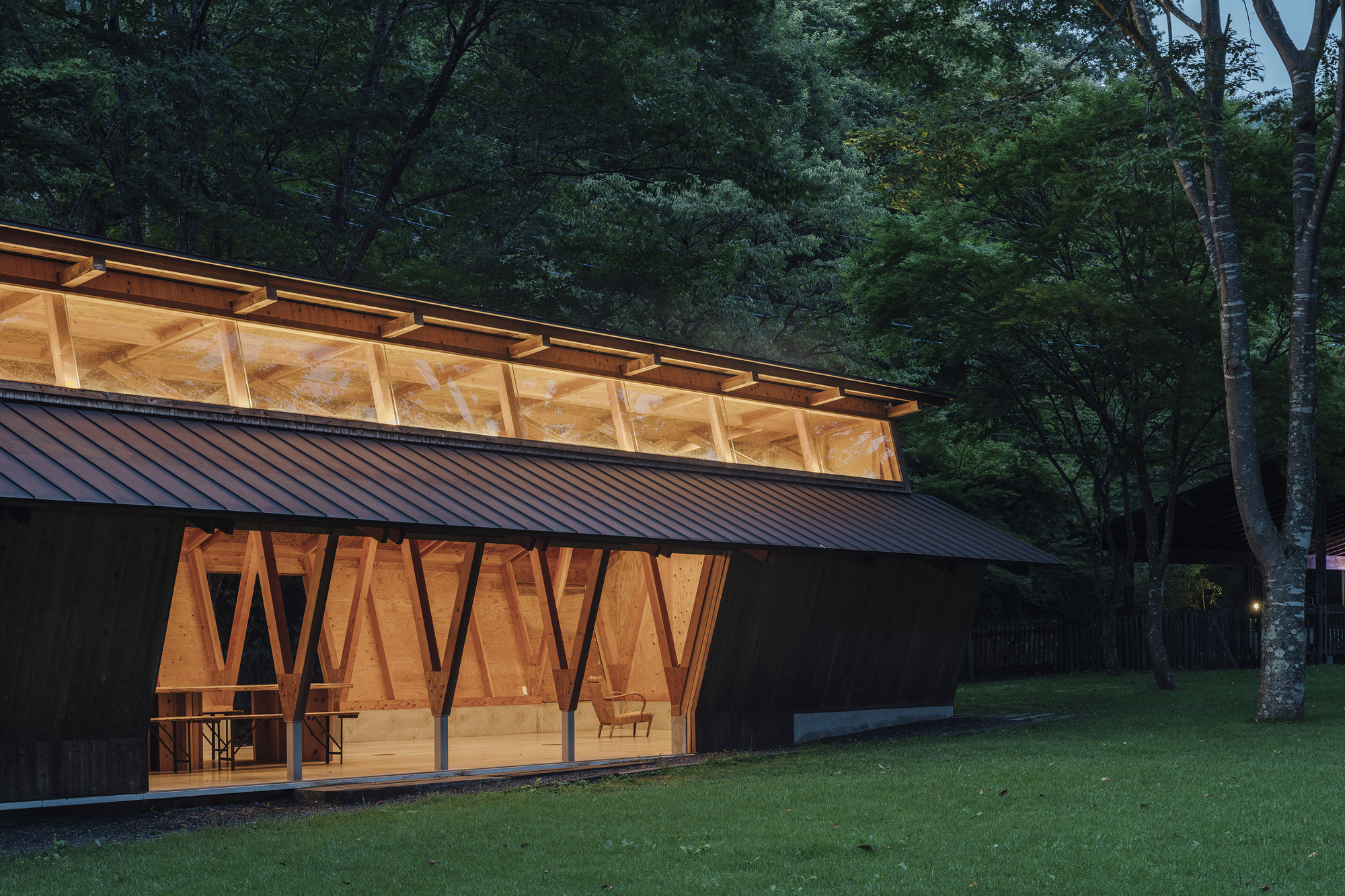 Diagonal braced timber facade with metal standing-seam roof and clerestory band illuminated at twilight