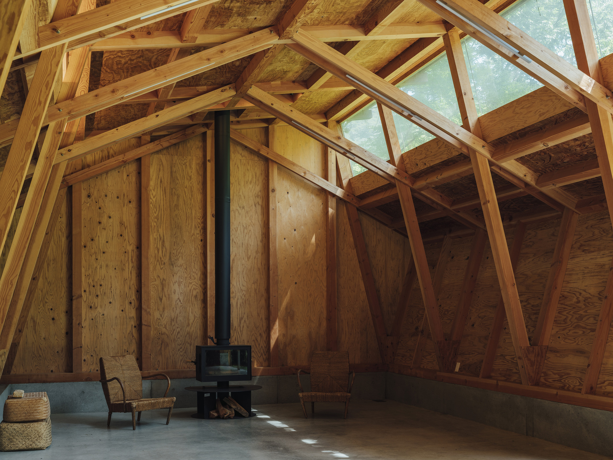 Plywood-clad interior wall with a wood stove on the concrete floor beneath angled timber trusses