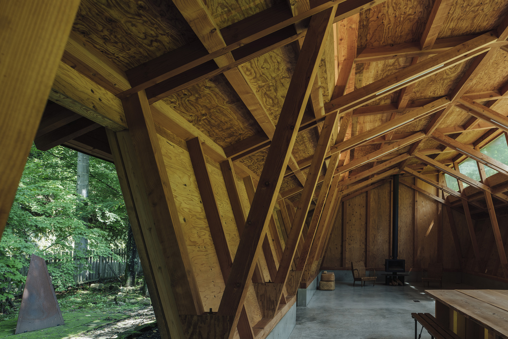 View through diagonal timber portal frames toward the forest and concrete floor in dappled light