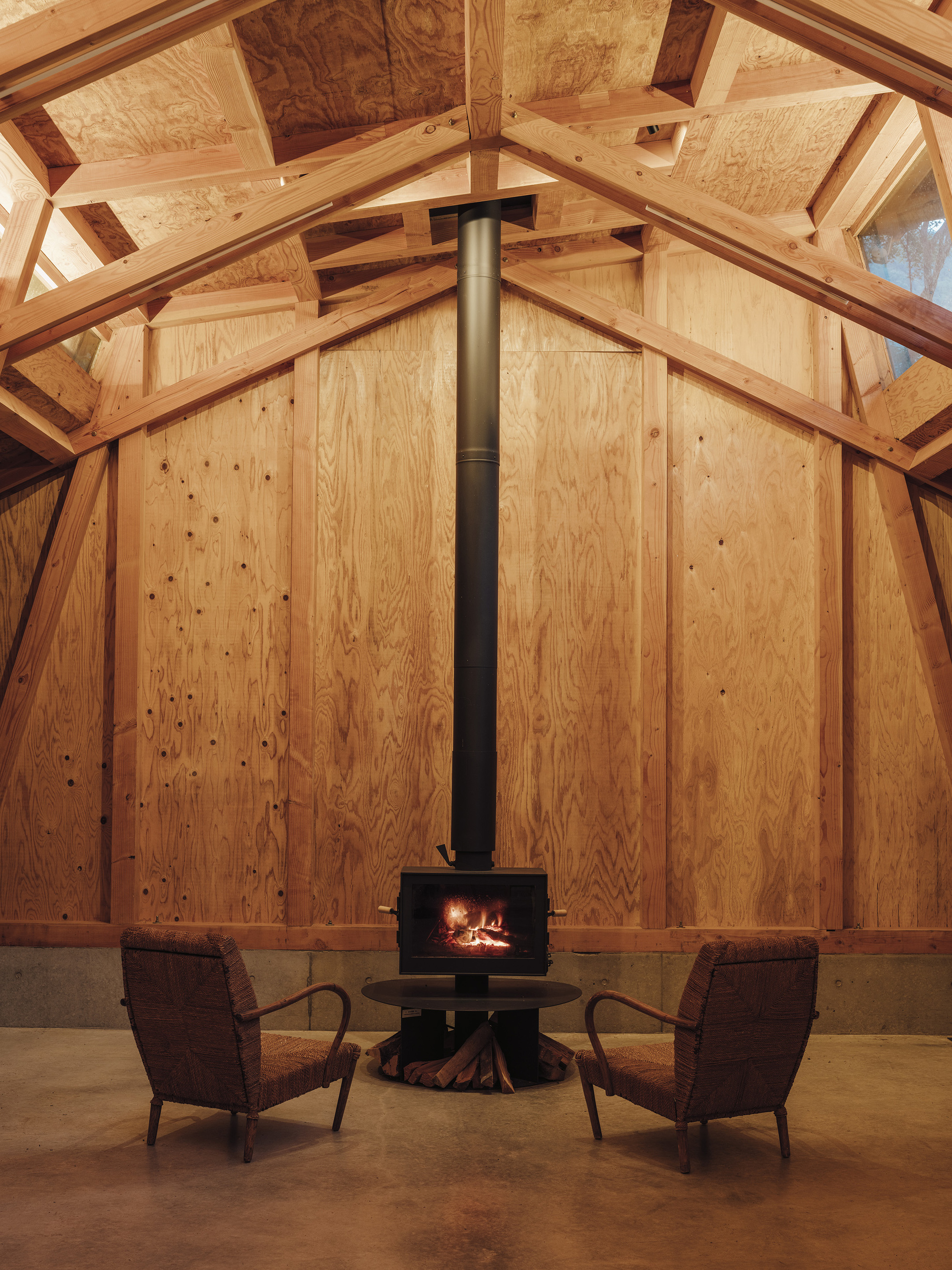 Freestanding wood stove with vertical flue pipe centered between two chairs under the vaulted timber ceiling